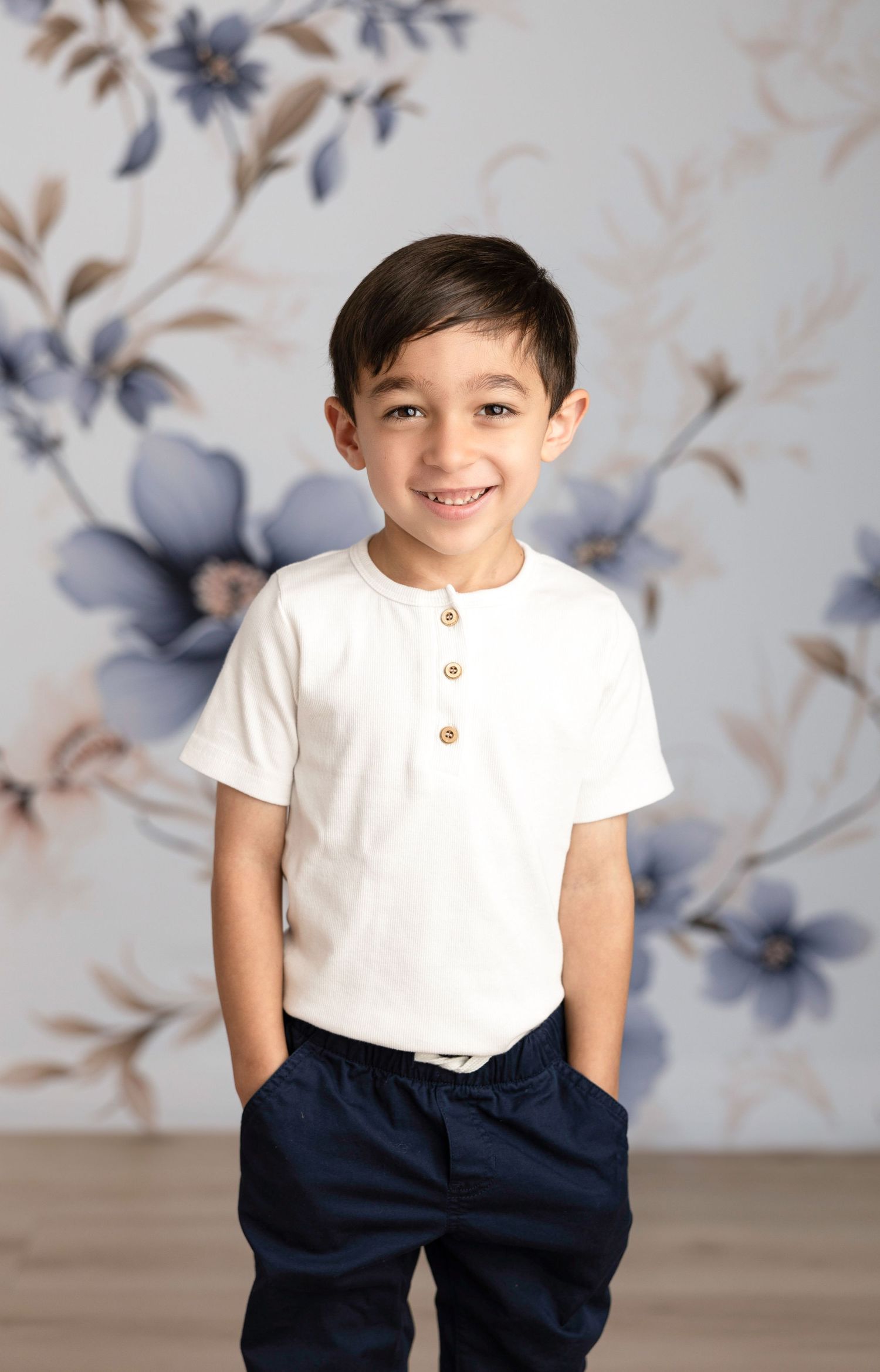 Young child in white henley shirt and navy pants poses against floral wallpaper.
