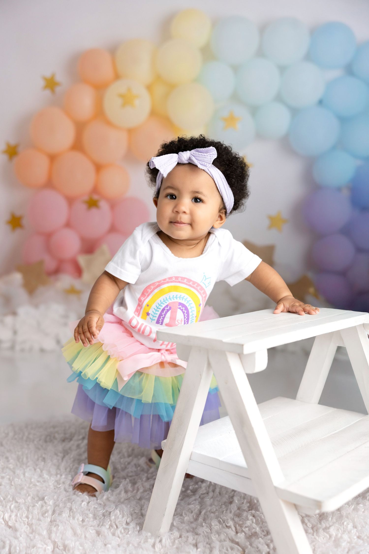 Baby in rainbow tutu dress sits near white bench with pastel balloon arch backdrop.