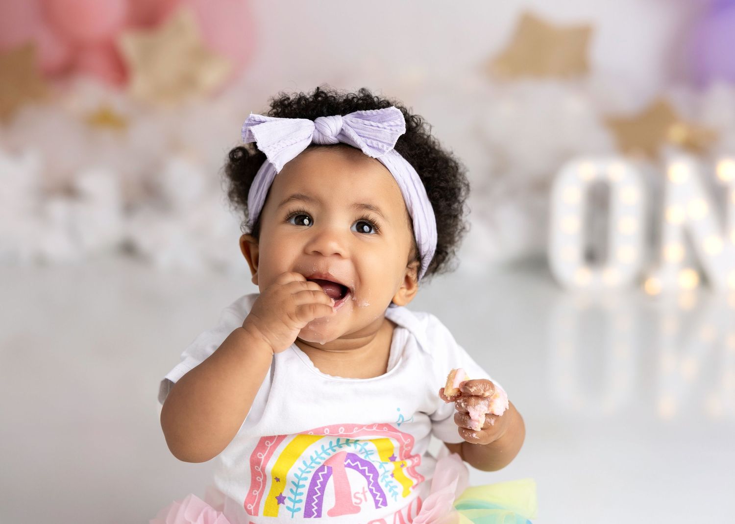Baby wearing rainbow outfit and bow explores festive birthday setup with pastel balloons.
