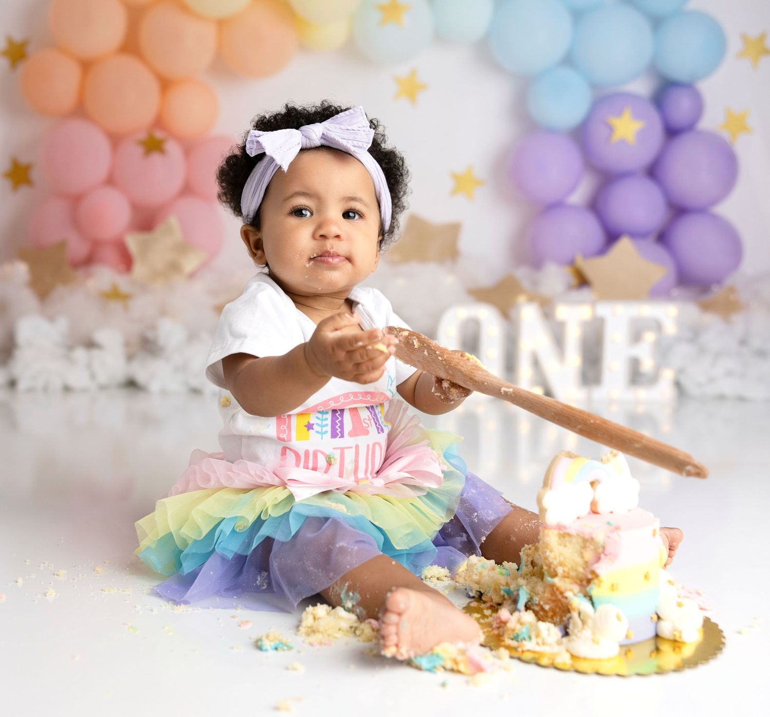 Baby in rainbow tutu dress enjoys cake smash photo session with pastel balloon decorations.