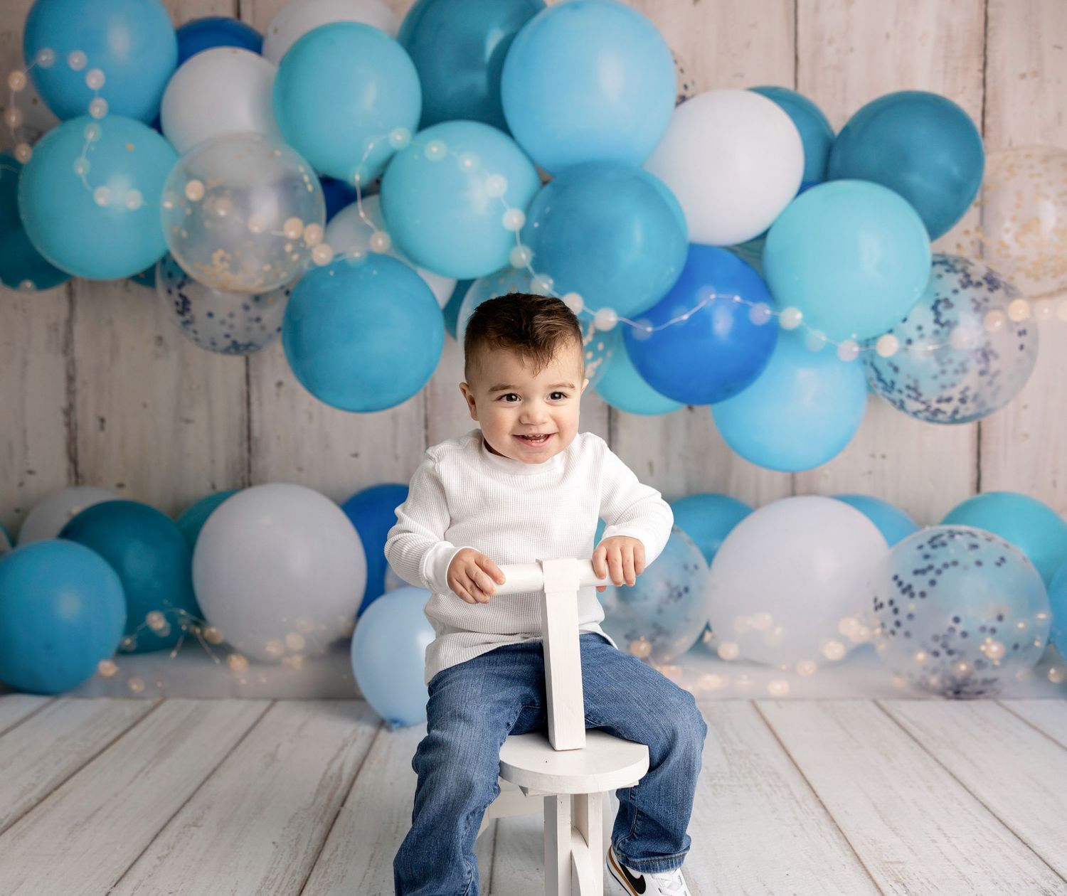 Child sitting on white chair with festive blue and white balloon arch background.
