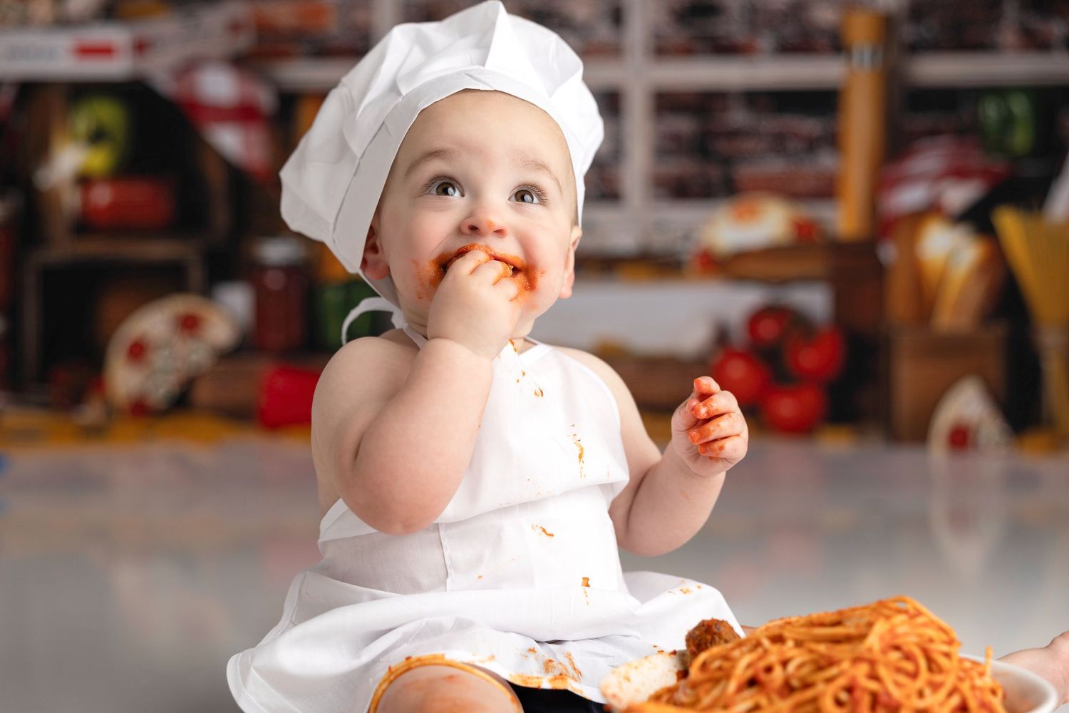 Baby in white chef outfit eating spaghetti with messy hands.