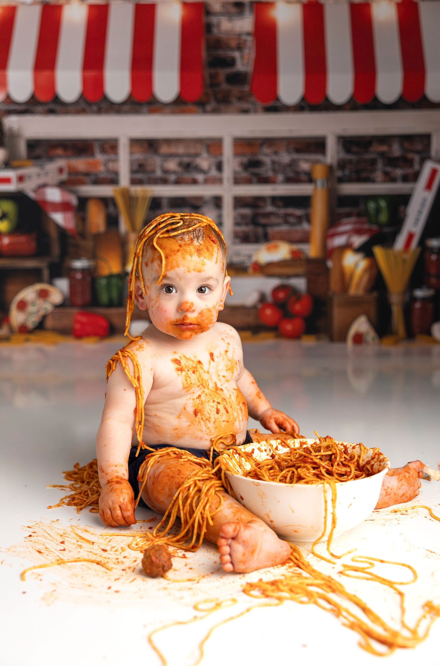 Messy baby covered in spaghetti sauce sitting with bowl of pasta.