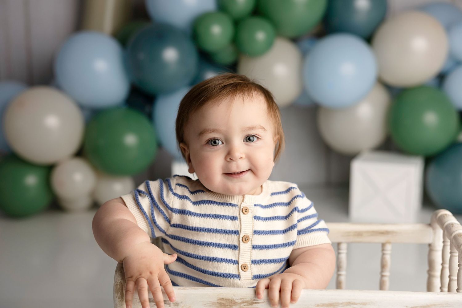 Smiling baby boy in striped outfit standing in white crib surrounded by blue, green, and white balloons.