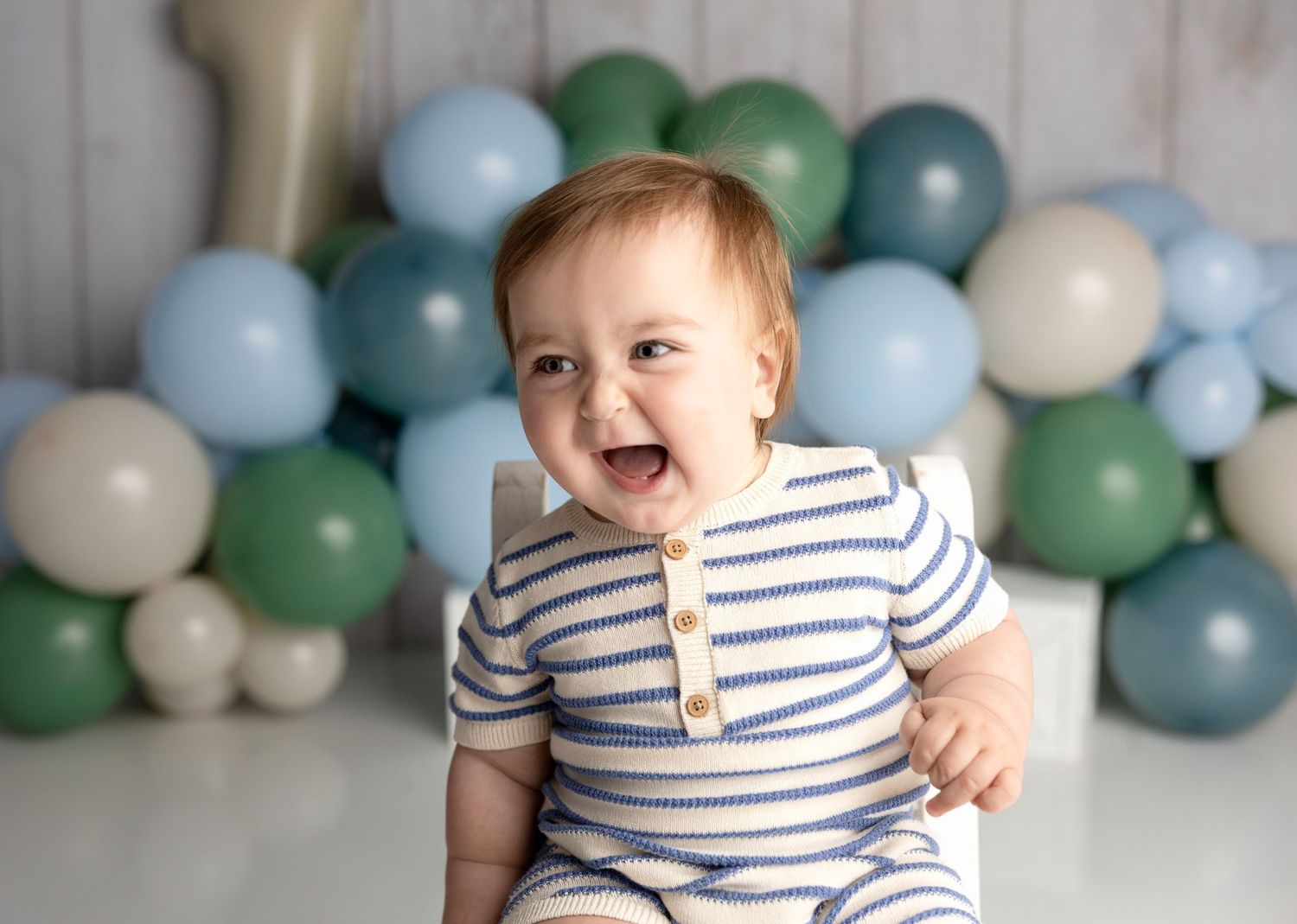 A joyful baby boy in a striped romper laughs during his first birthday cake smash with blue and green balloons.