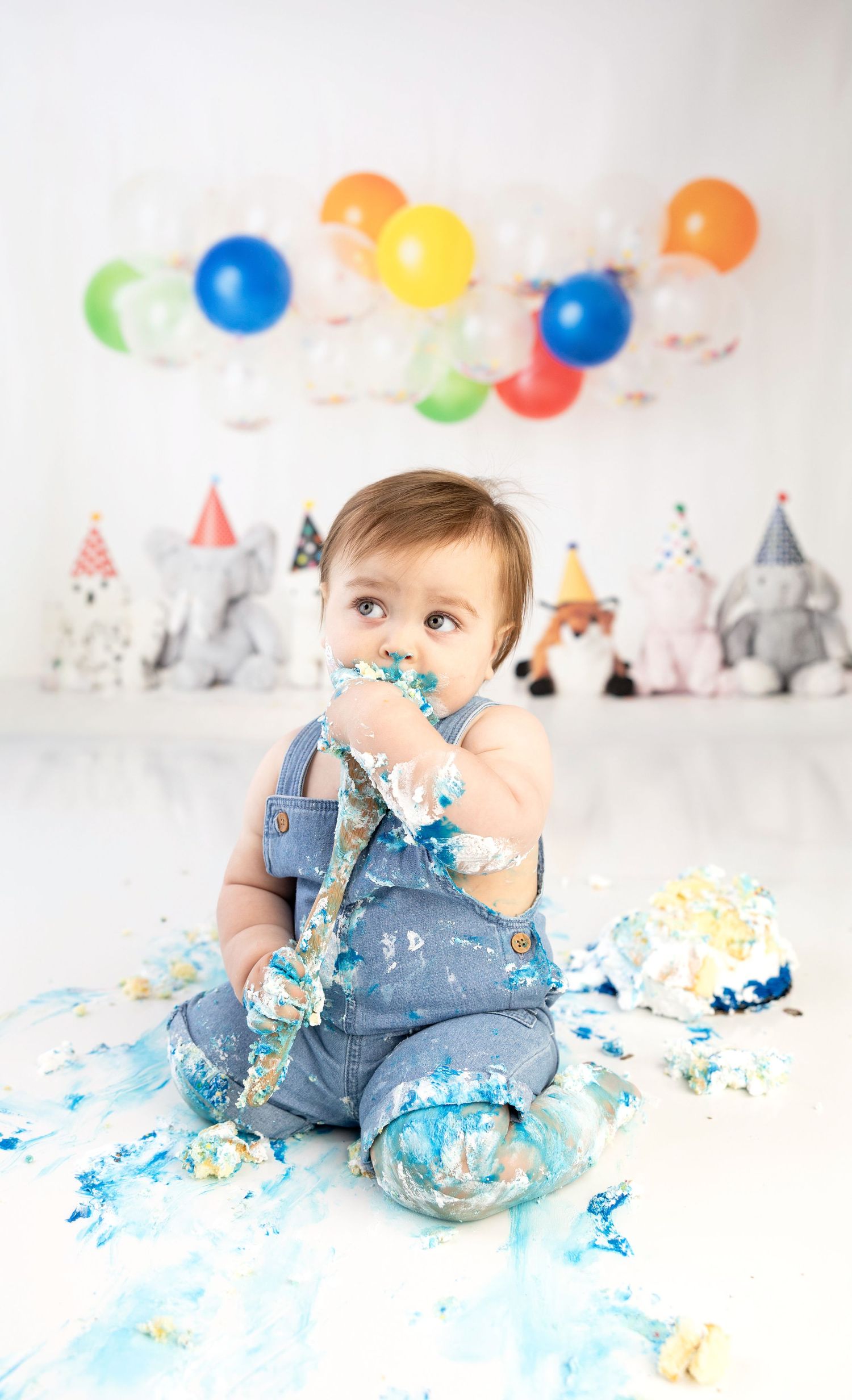 A baby in blue overalls smashes their first birthday cake surrounded by colorful balloons and party decorations.