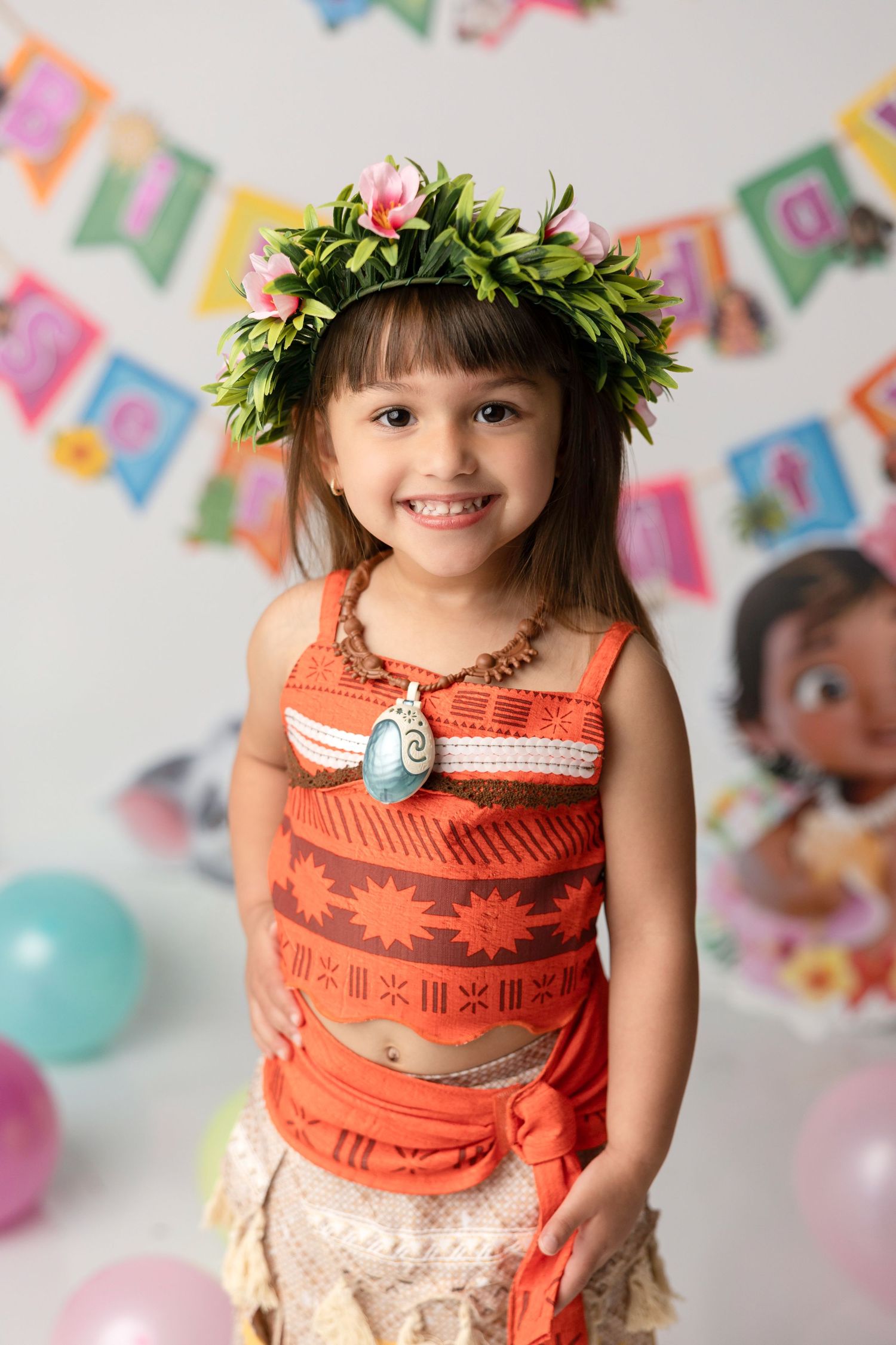Young girl in Moana costume with floral crown smiles at birthday party with colorful banners and balloons.