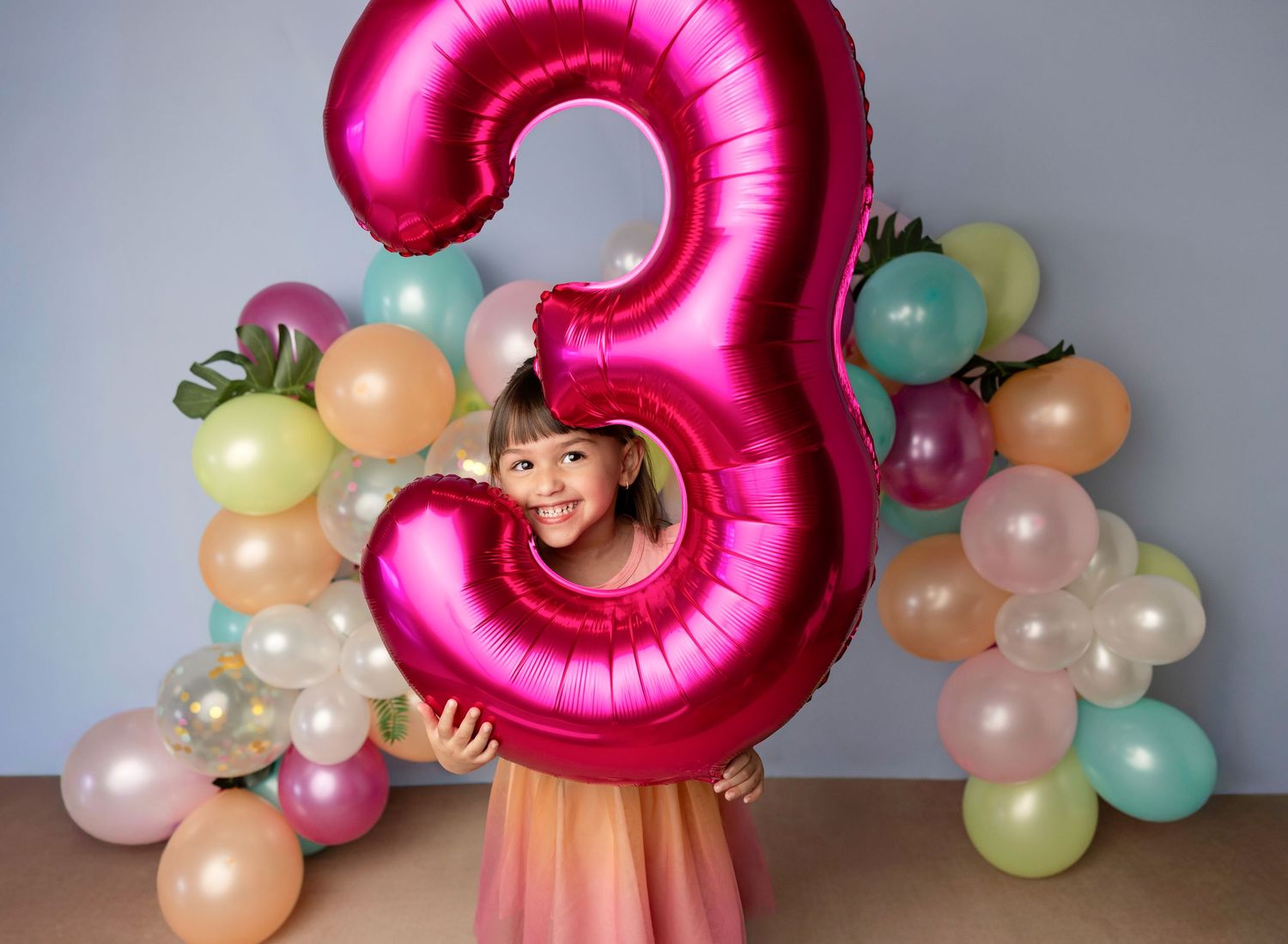 A young girl smiles through a giant pink number 3 balloon surrounded by colorful balloons at a birthday party.