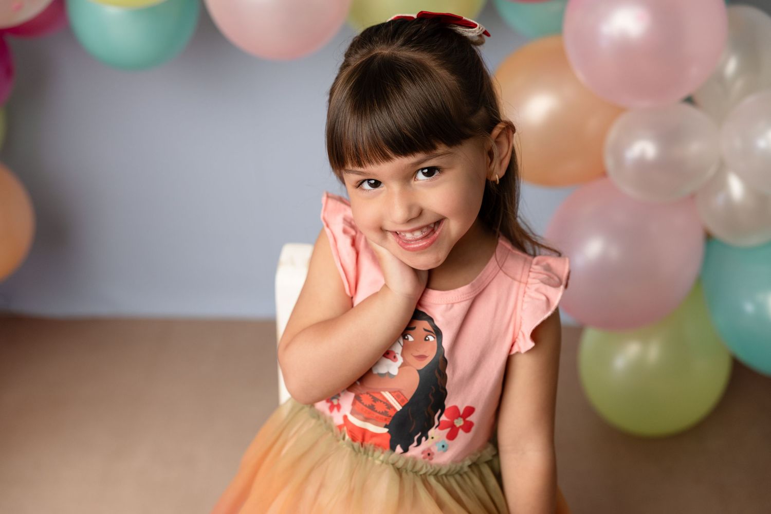 Young girl in pink Moana dress smiling at birthday party with colorful balloons in background.