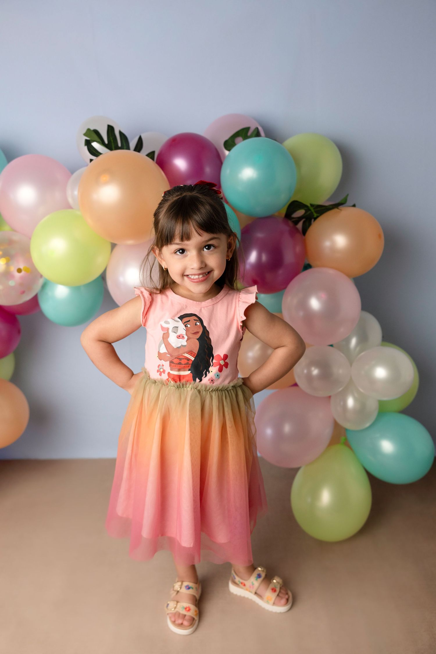 A smiling little girl in a colorful Moana dress poses with hands on hips in front of a vibrant balloon wall.