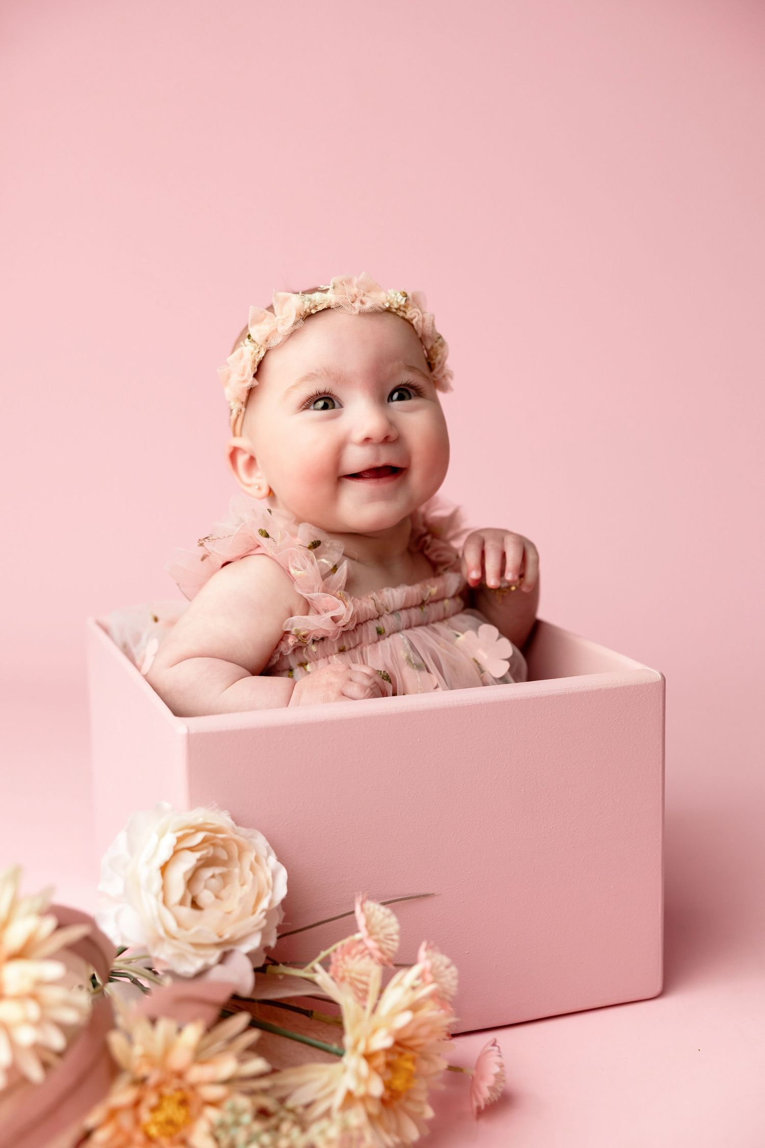 Smiling baby girl in pink ruffled dress and floral headband sitting in a pink box with flowers.