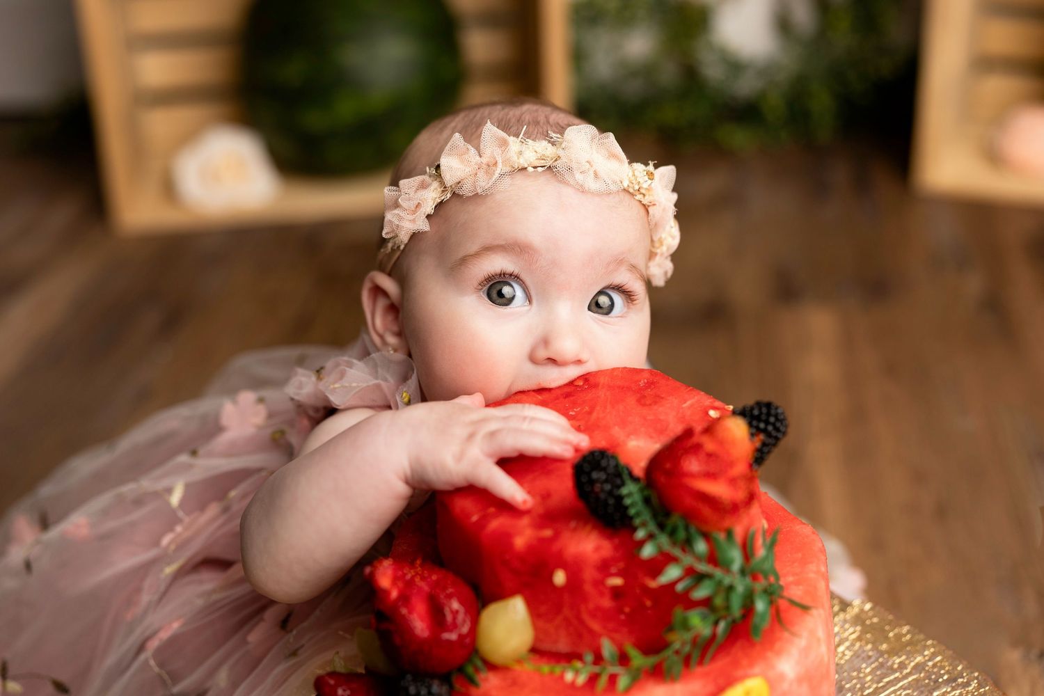Baby girl in pink tutu and floral headband smiling while touching a fruit-decorated cake at her first birthday.