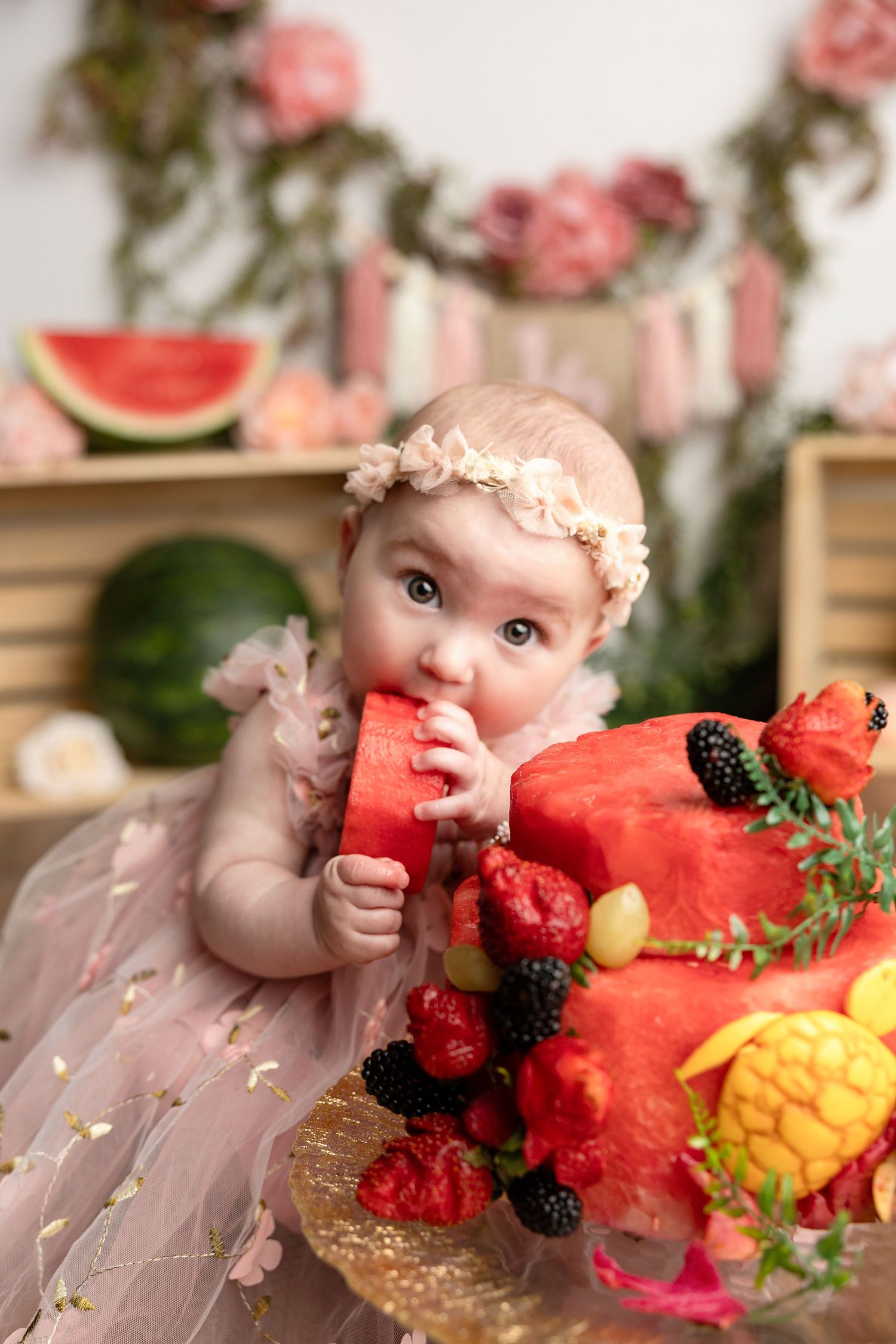 Baby girl in pink tutu eating watermelon slice beside a fruit-decorated cake at her first birthday celebration.