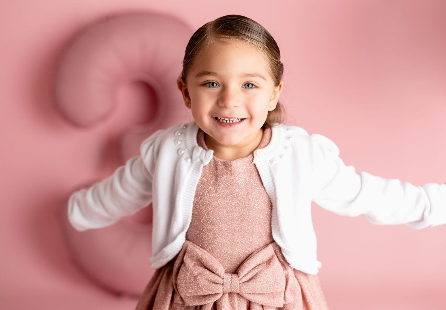 Joyful portrait in white cardigan and pink dress against pink background.