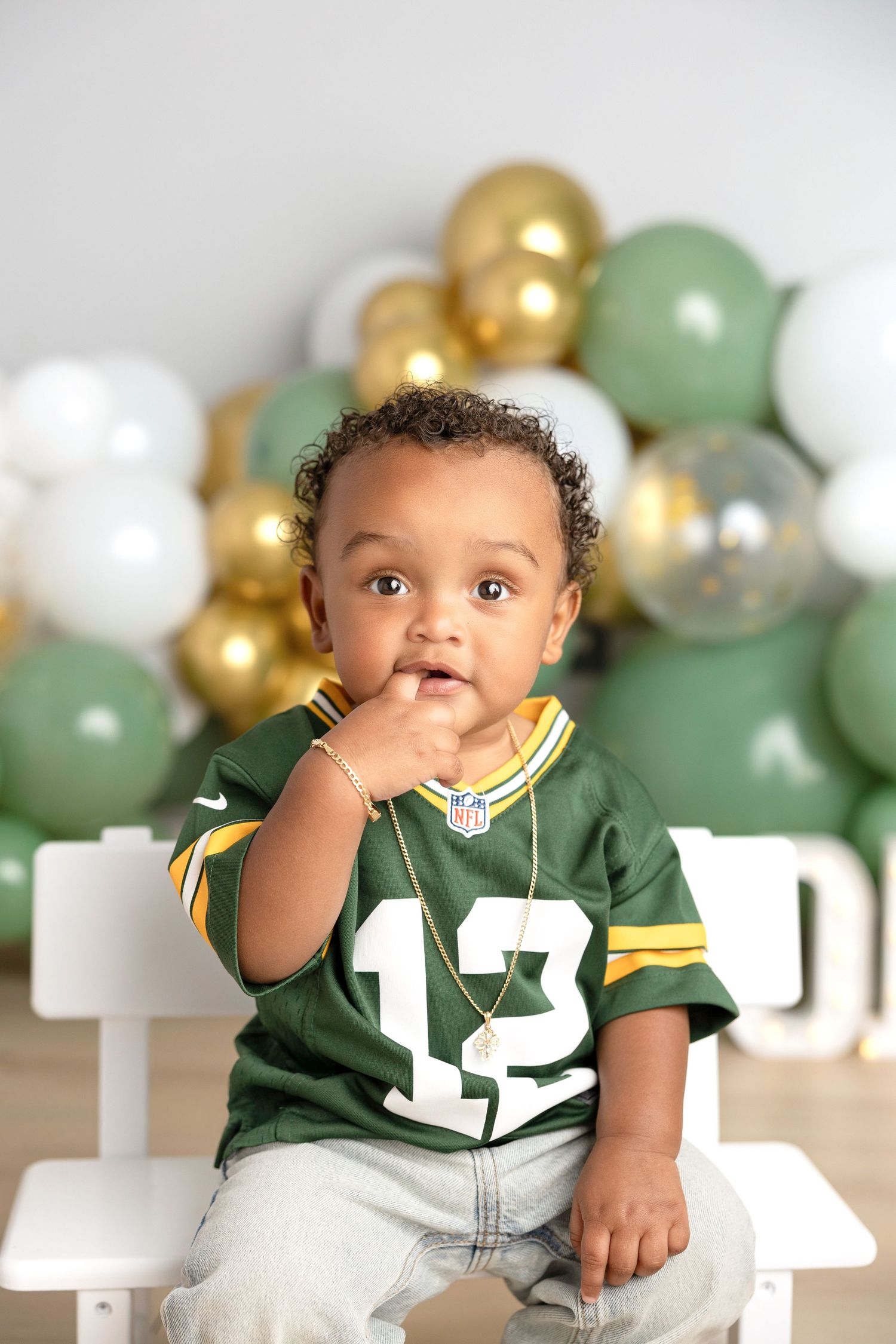 Baby in green sports jersey sitting among gold and sage balloon decorations.