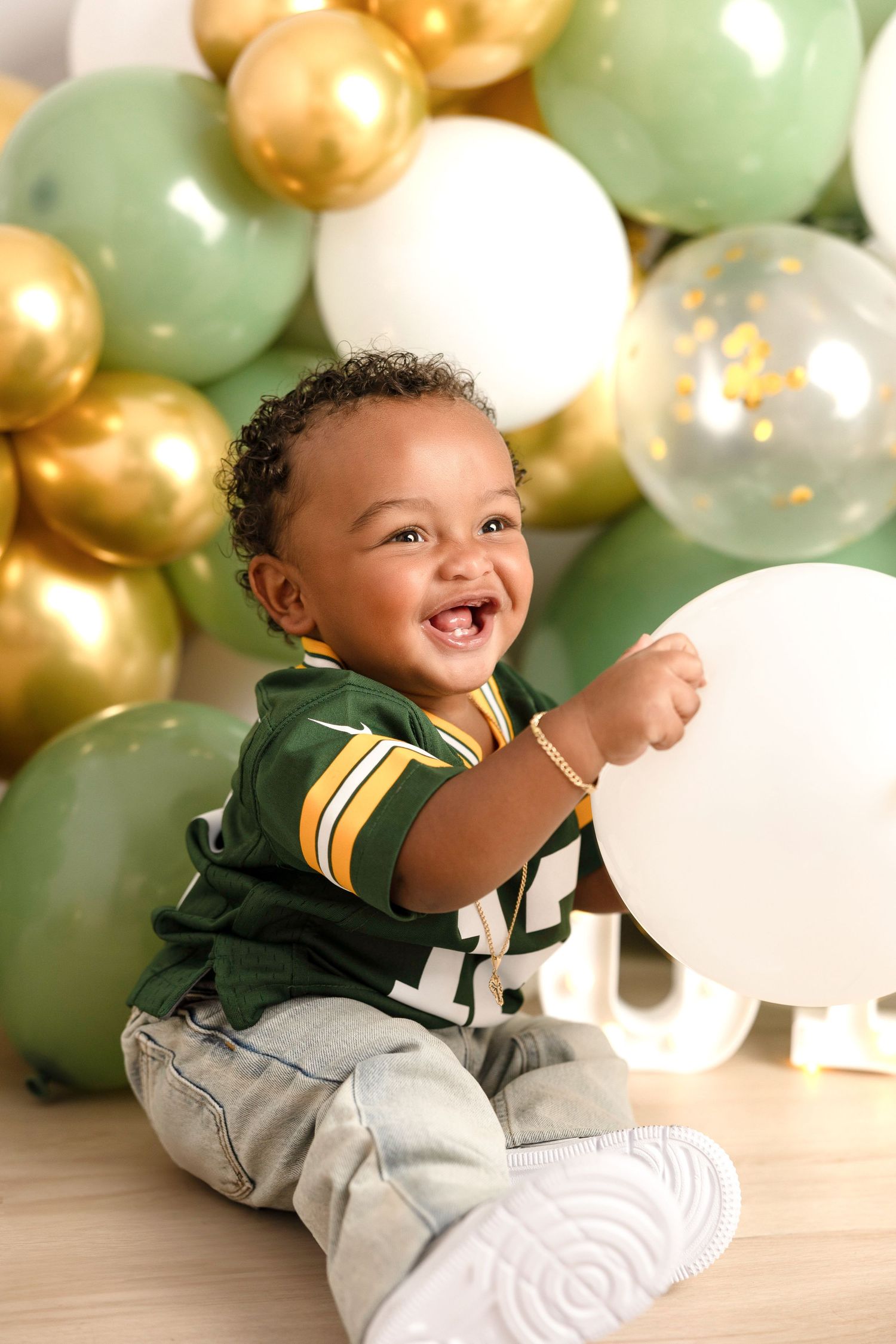 Joyful baby in sports attire playing among festive gold and green balloons.
