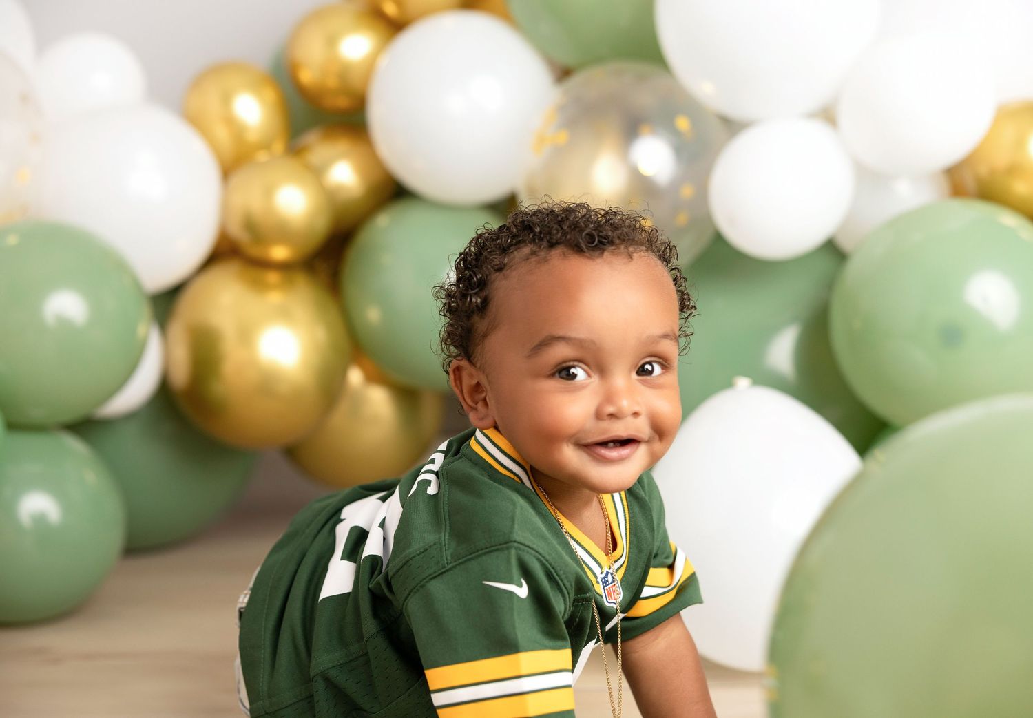 Baby wearing green and white sports jersey smiles against balloon backdrop.