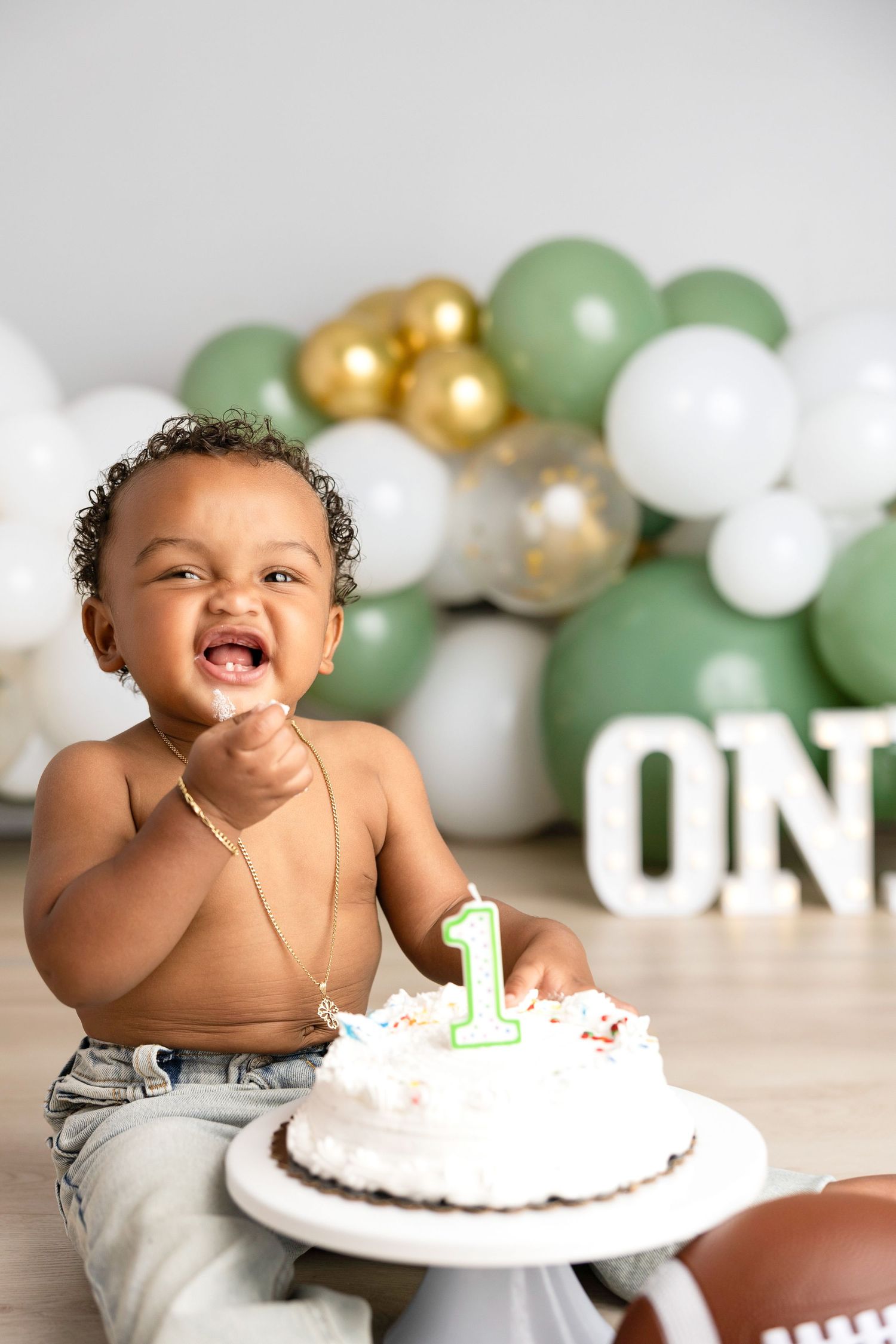 Baby in diaper celebrates first birthday with white cake and balloon decorations.