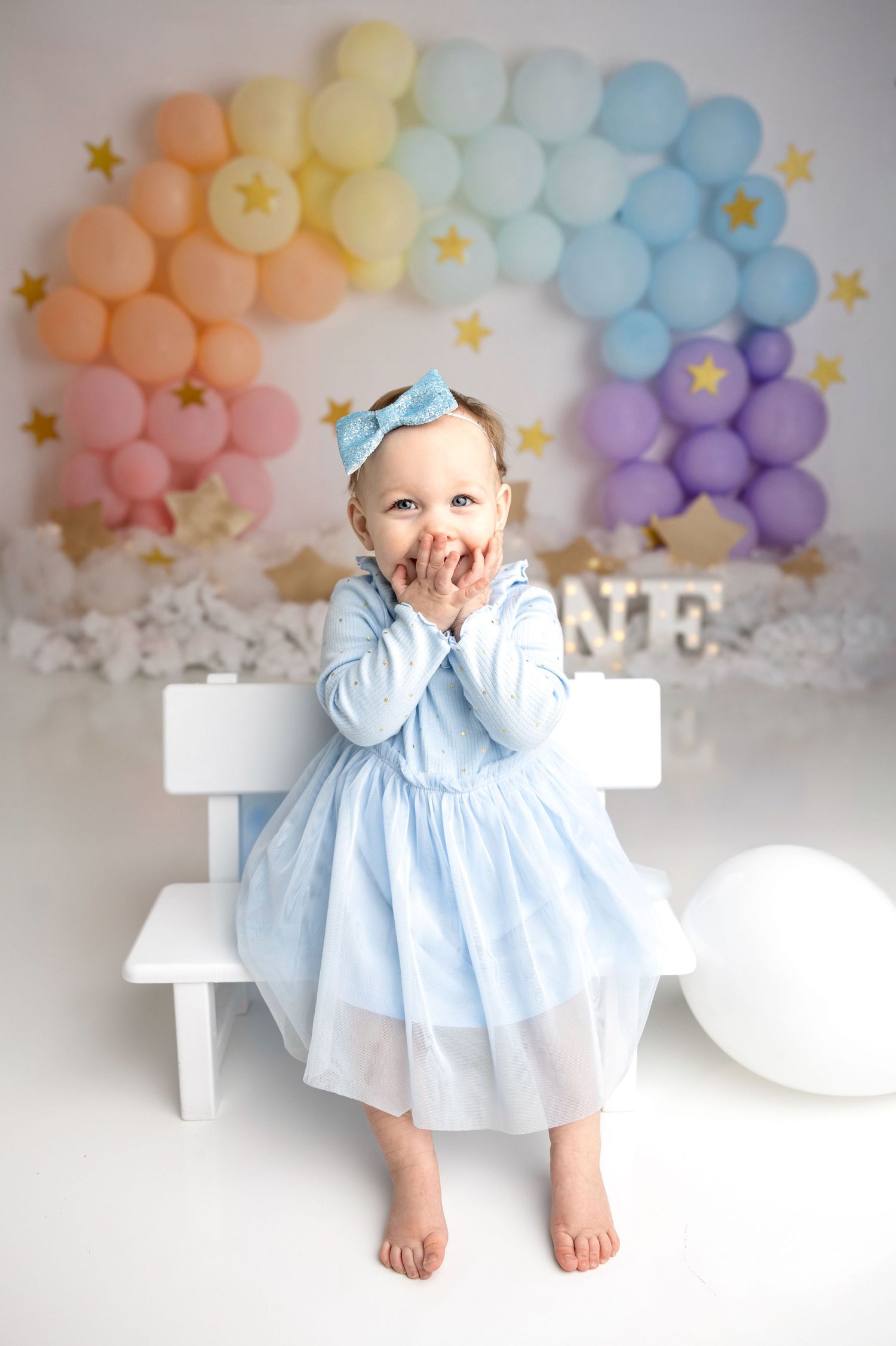 Baby in blue dress sitting on white bench with rainbow balloon arch background.