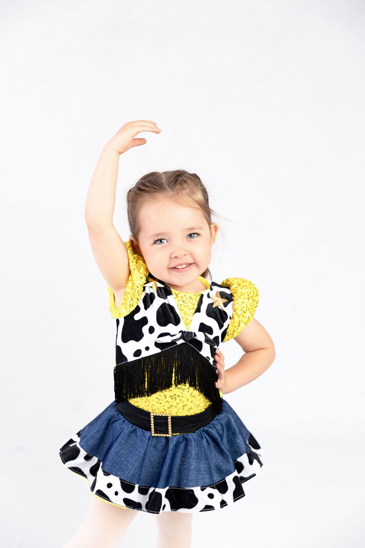 Smiling toddler girl in cowgirl dance costume with cow print, yellow sequins, and denim skirt, posing with arm raised.