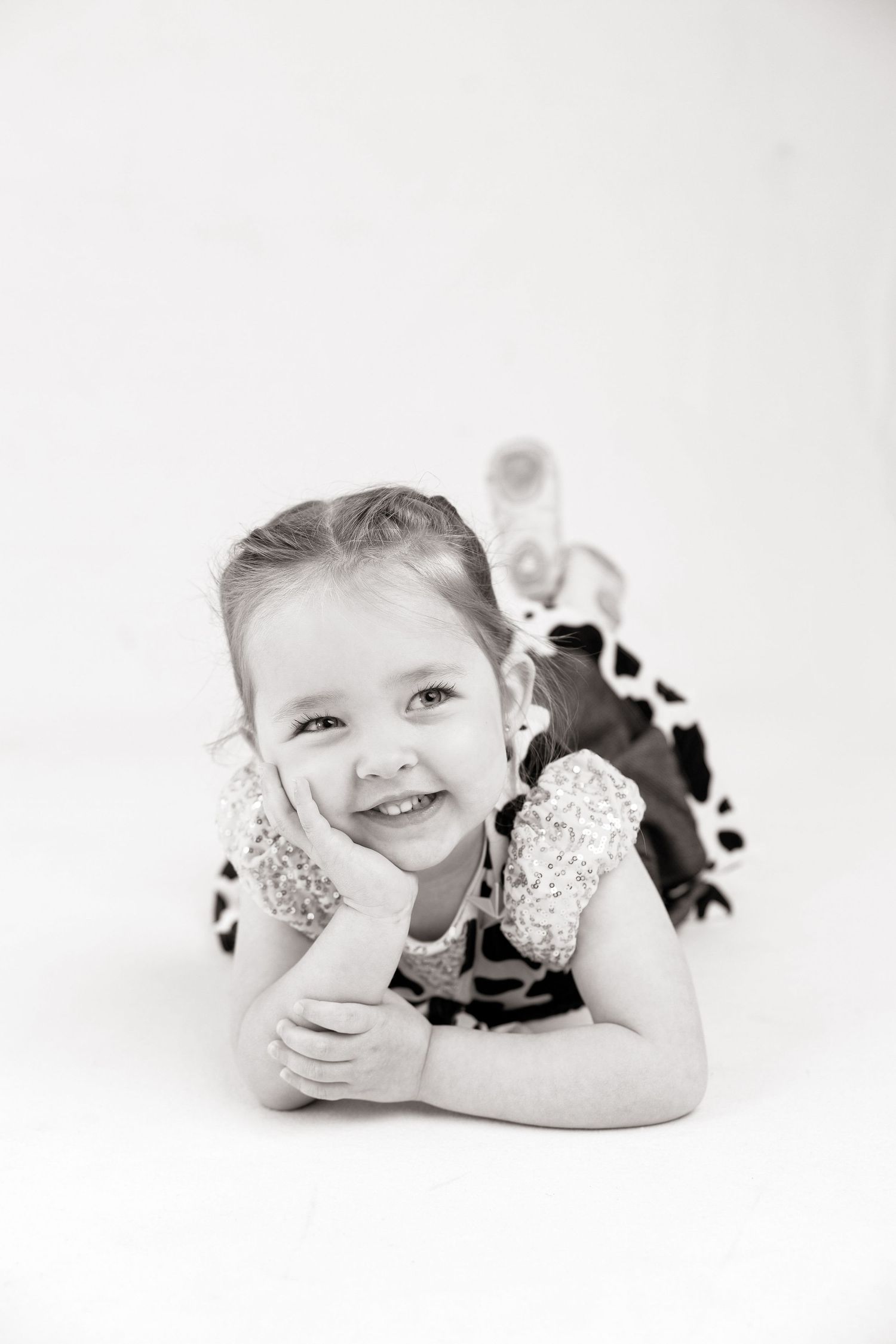 Black and white photo of a smiling young girl lying on floor, resting chin on hand, wearing a cow print dress.