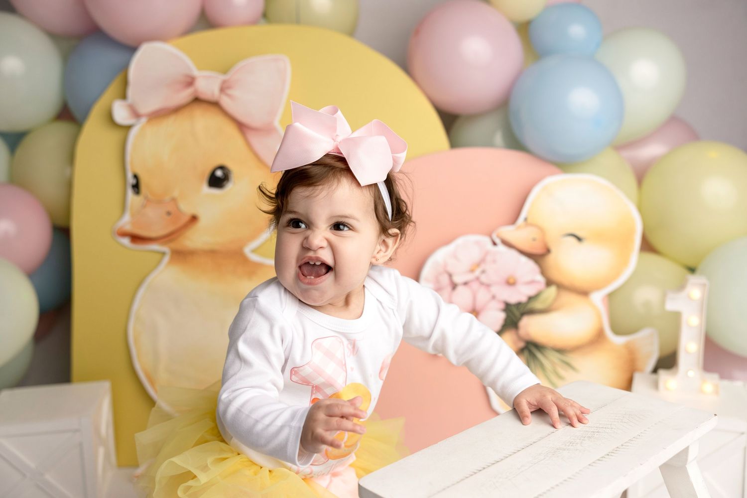 Baby girl in yellow tutu smiles at first birthday party with duck theme balloons and decorations.