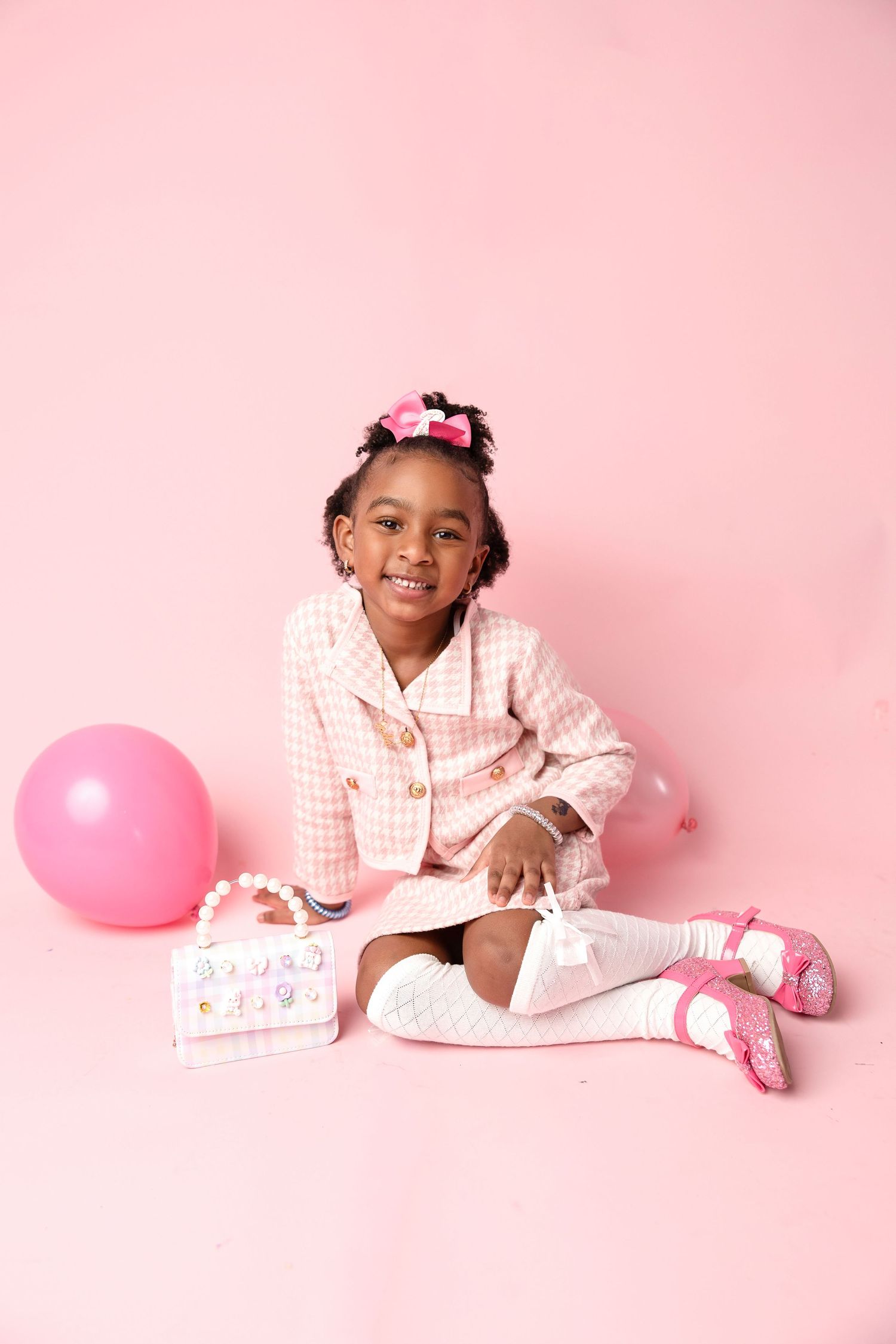 A child in white pajamas poses playfully with pink balloons against a pink background for a studio photoshoot.