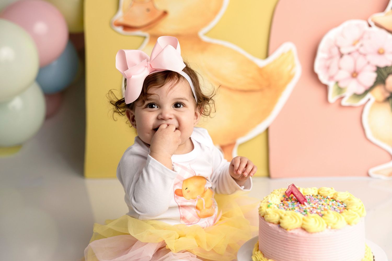 Baby girl in yellow tutu and pink bow celebrates first birthday with yellow cake at rubber duck themed party.