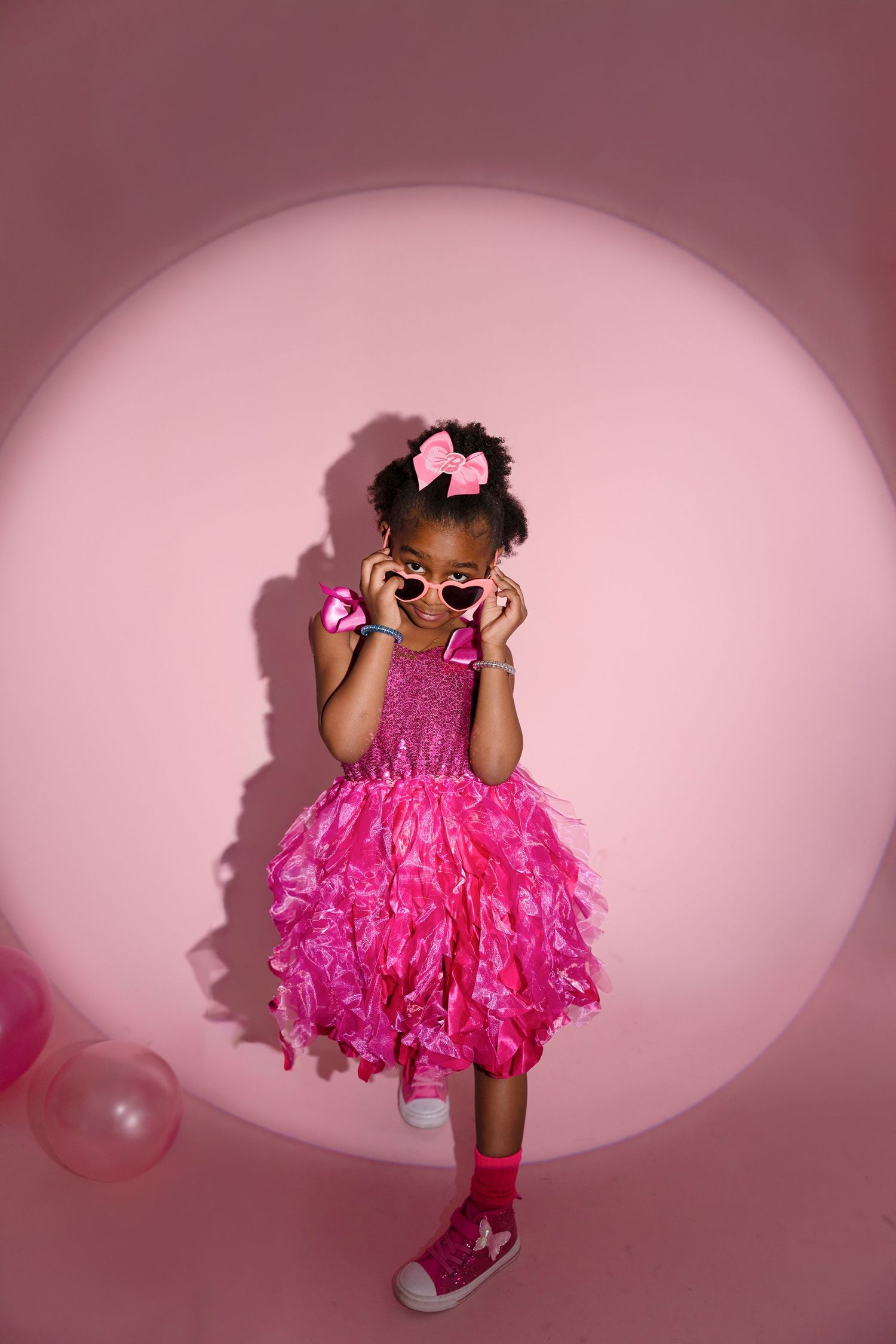 A series of four photos showing a child in a bright pink tulle dress and sunglasses posing playfully against a pink backdrop.