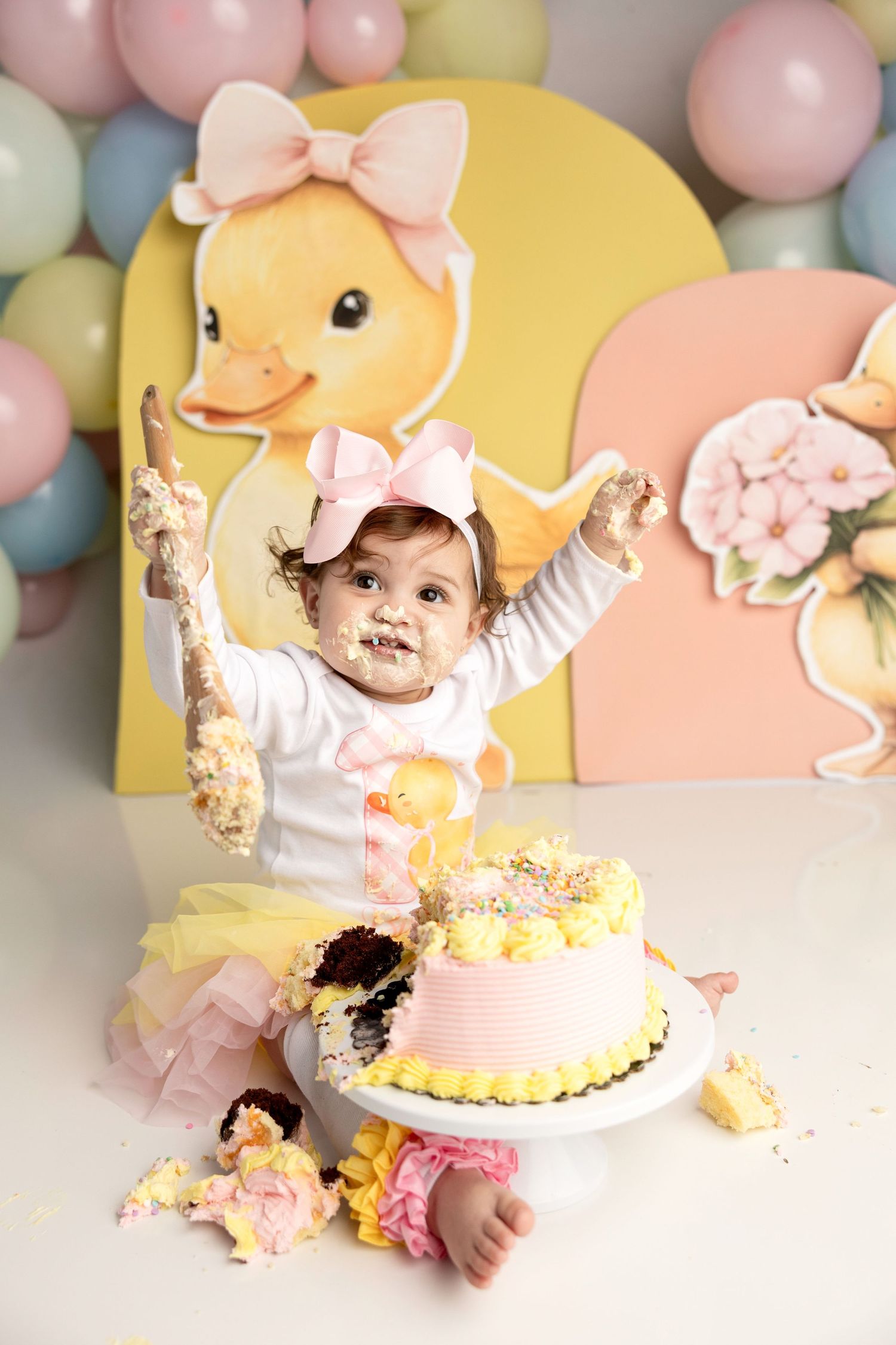 Baby girl celebrates first birthday smashing yellow and pink cake, with duck themed balloons and decorations in background.