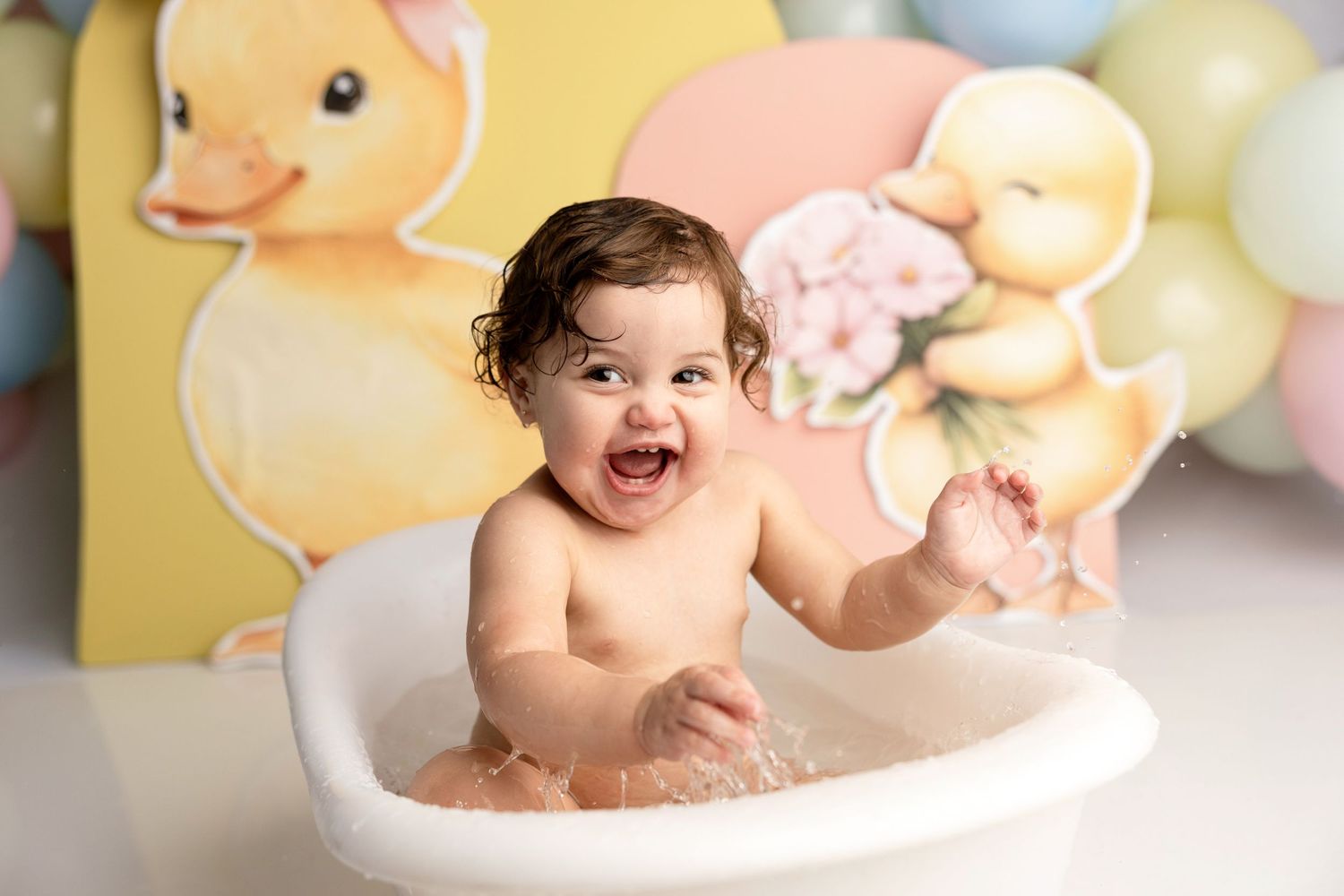 Happy baby laughing in a white bathtub during a rubber duck themed photo shoot with pastel balloons.