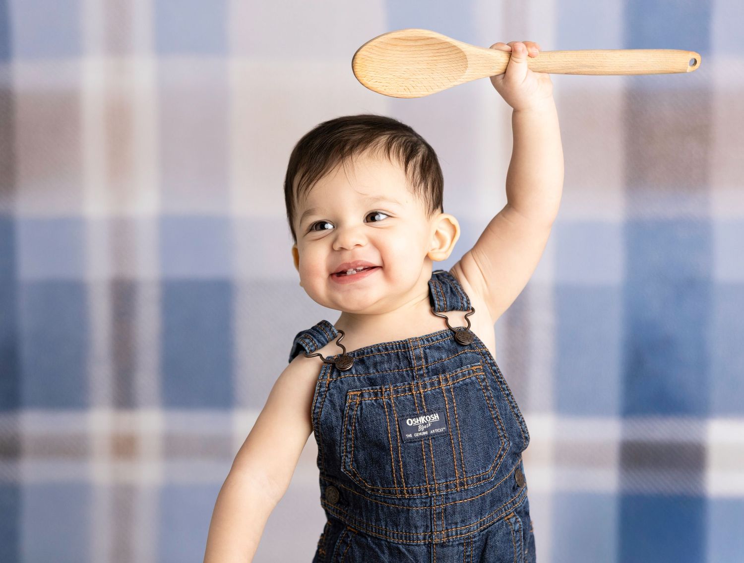 Baby in denim overalls playing with wooden spoon against blue backdrop.