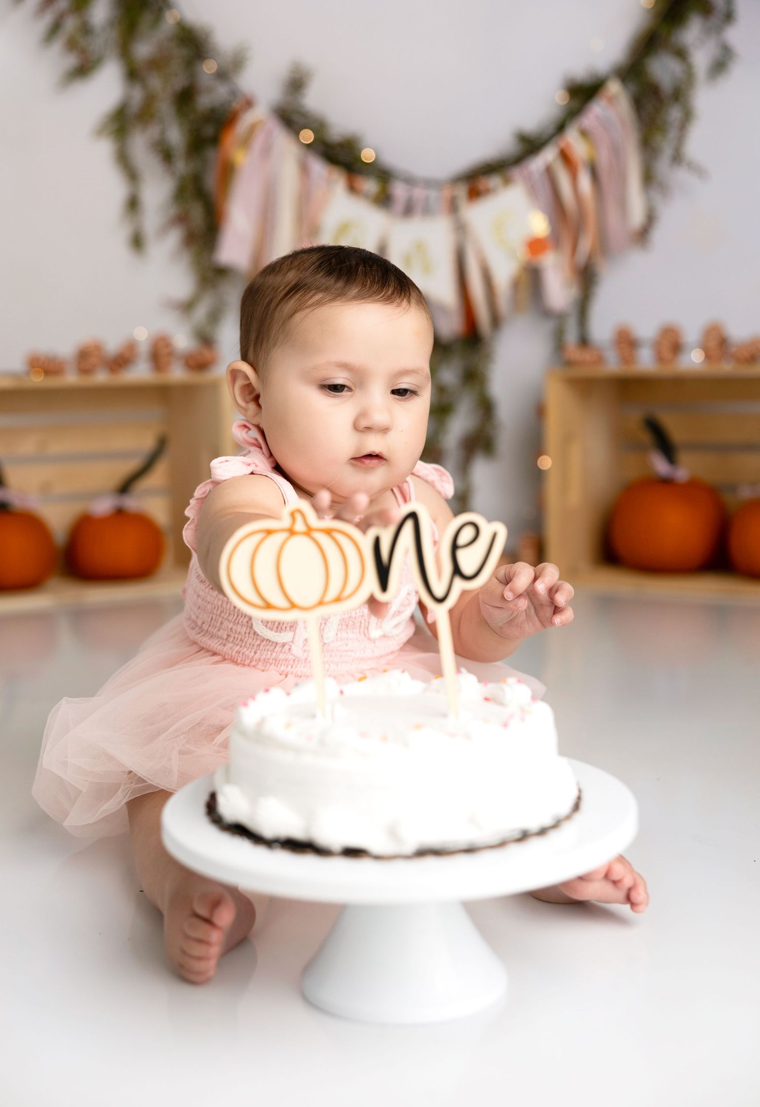 Baby's first cake smash photos with pink dress and pumpkin themed cake on white table.