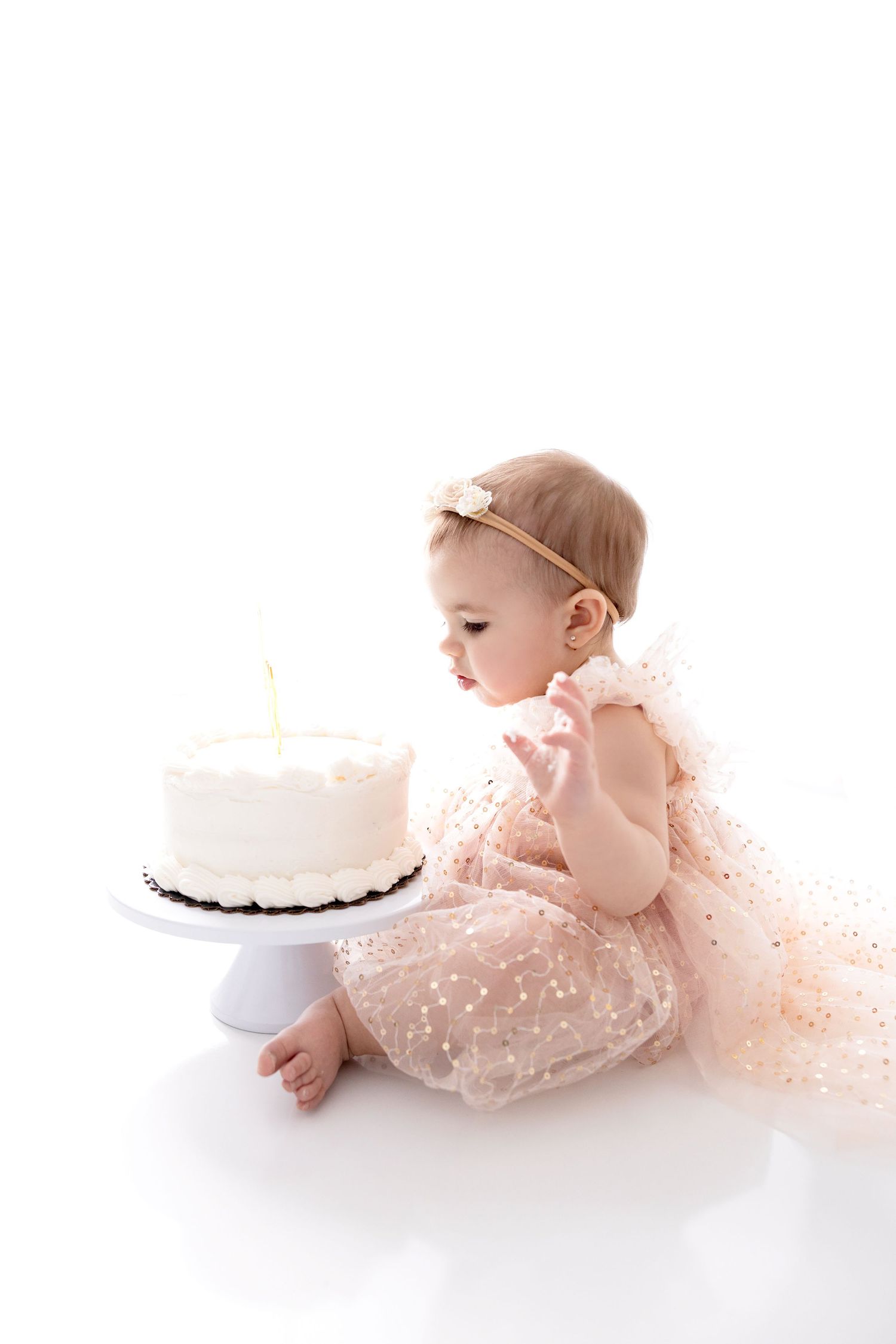 Baby in pink tulle dress crawls toward white cake on pedestal against white background in series of photos.