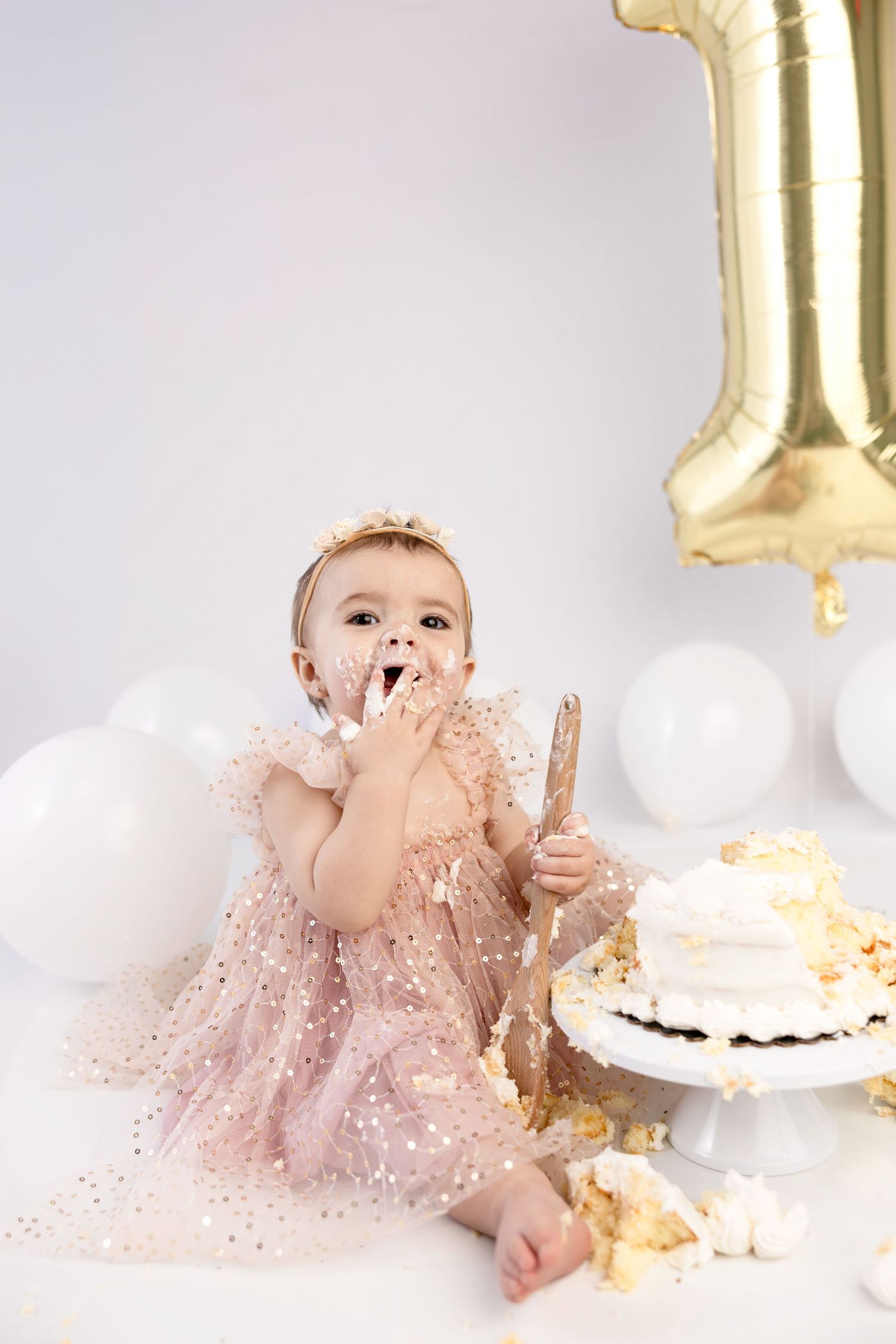 Baby in pink sparkly dress celebrates first birthday with smashed cake and gold balloon decorations.