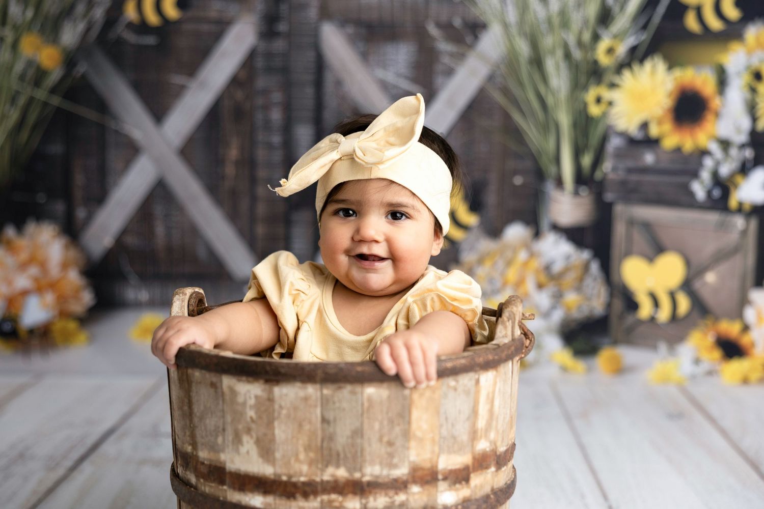 Baby sitting in rustic wooden bucket with bee decorations in background.