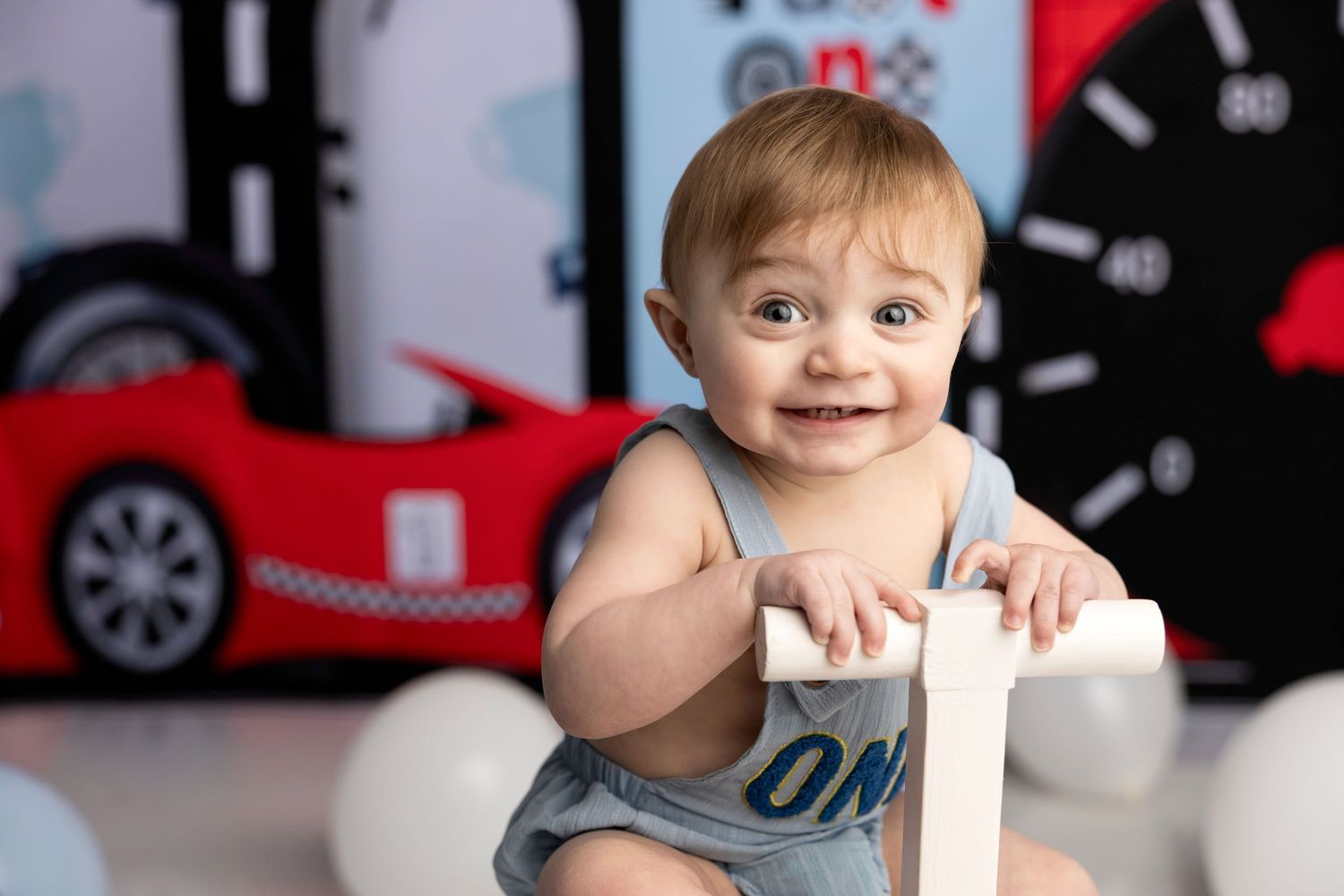 A smiling baby in a gray onesie sits in front of a red race car bed and decorative backdrop.