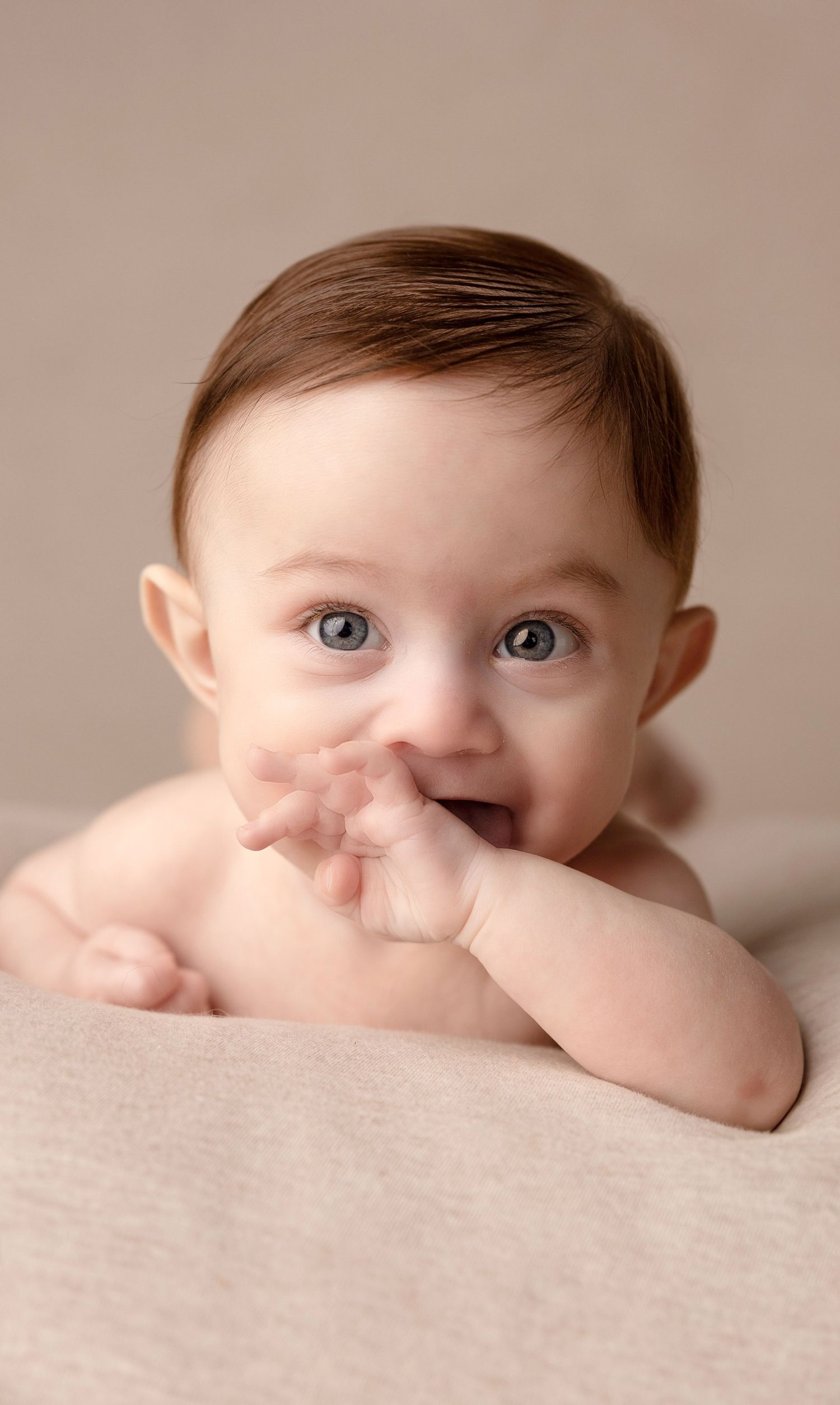Baby lying on beige blanket with wide eyes and hands near mouth.