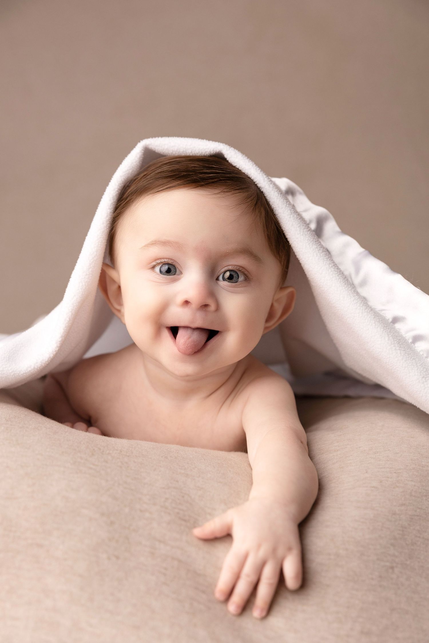 Baby peeks out from under white blanket with playful expression.