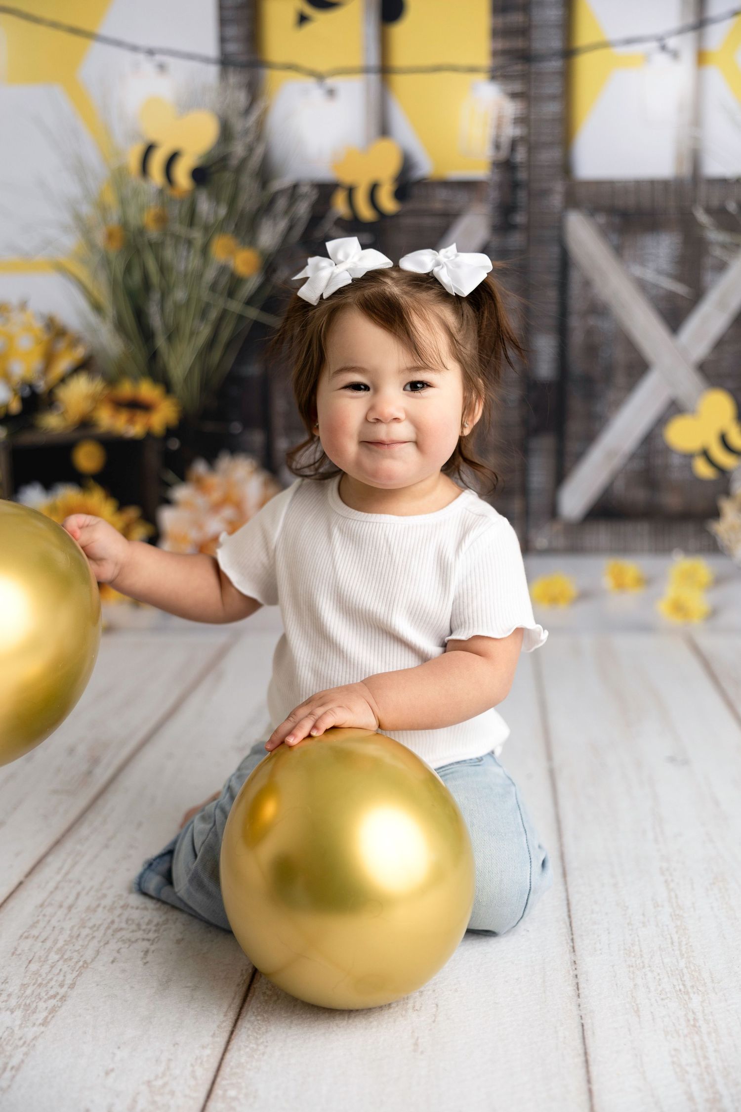 Baby in white shirt and jeans playing with golden balloon against bee-themed backdrop.