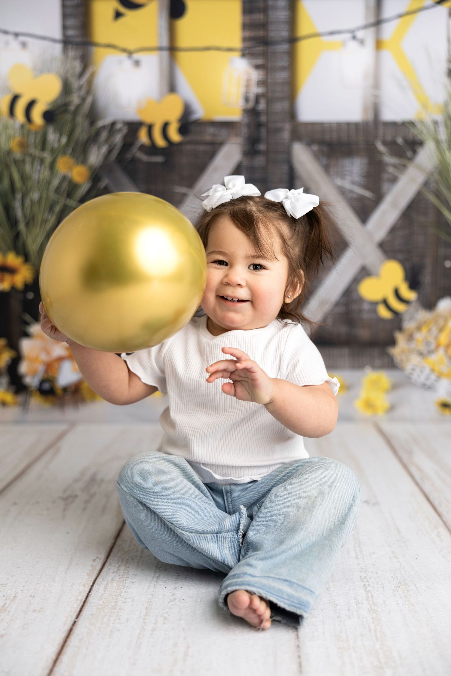 Baby in casual outfit playing with large golden balloon in bee-themed setting.