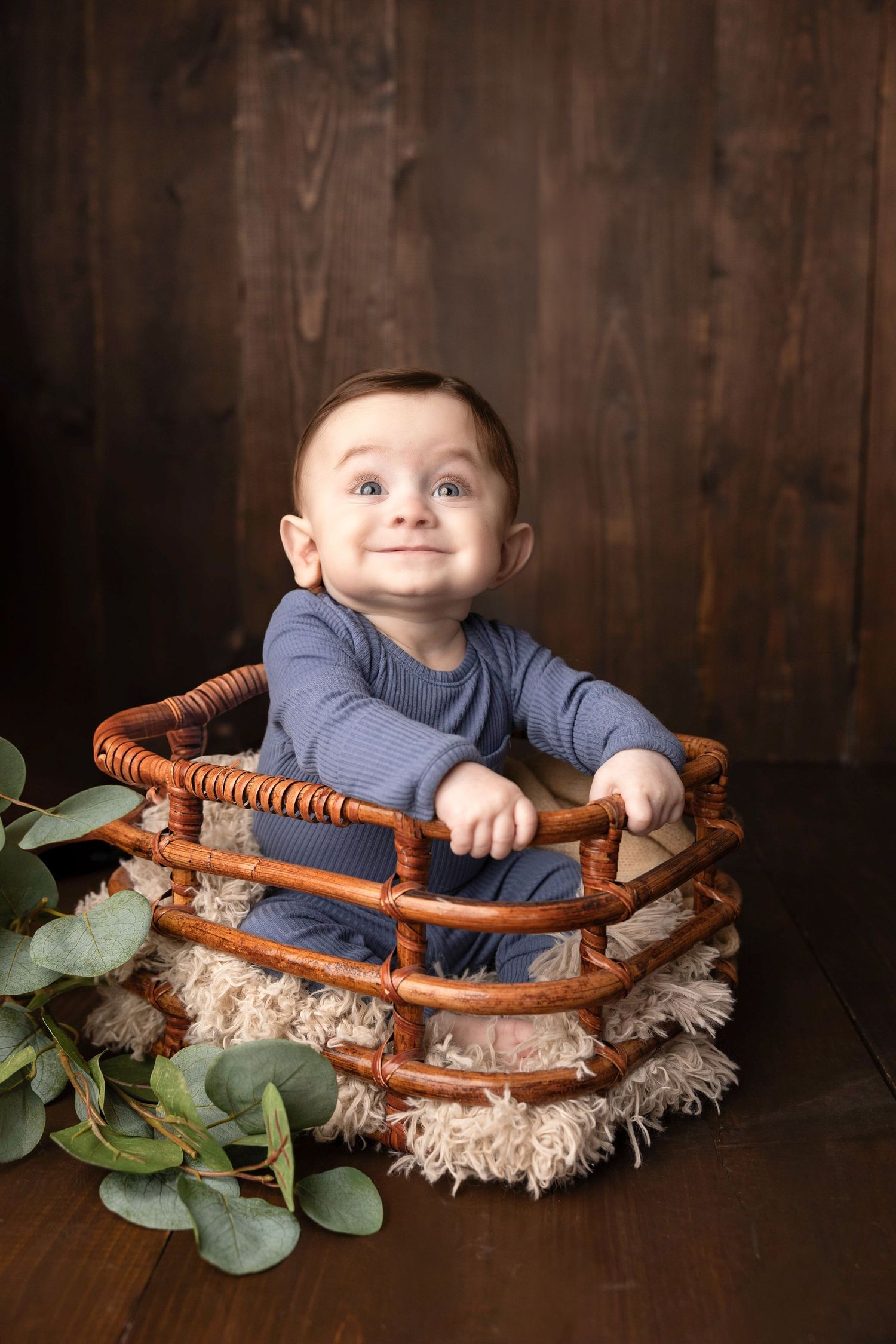 Baby in blue outfit sits in wooden basket with eucalyptus leaves and fur padding.