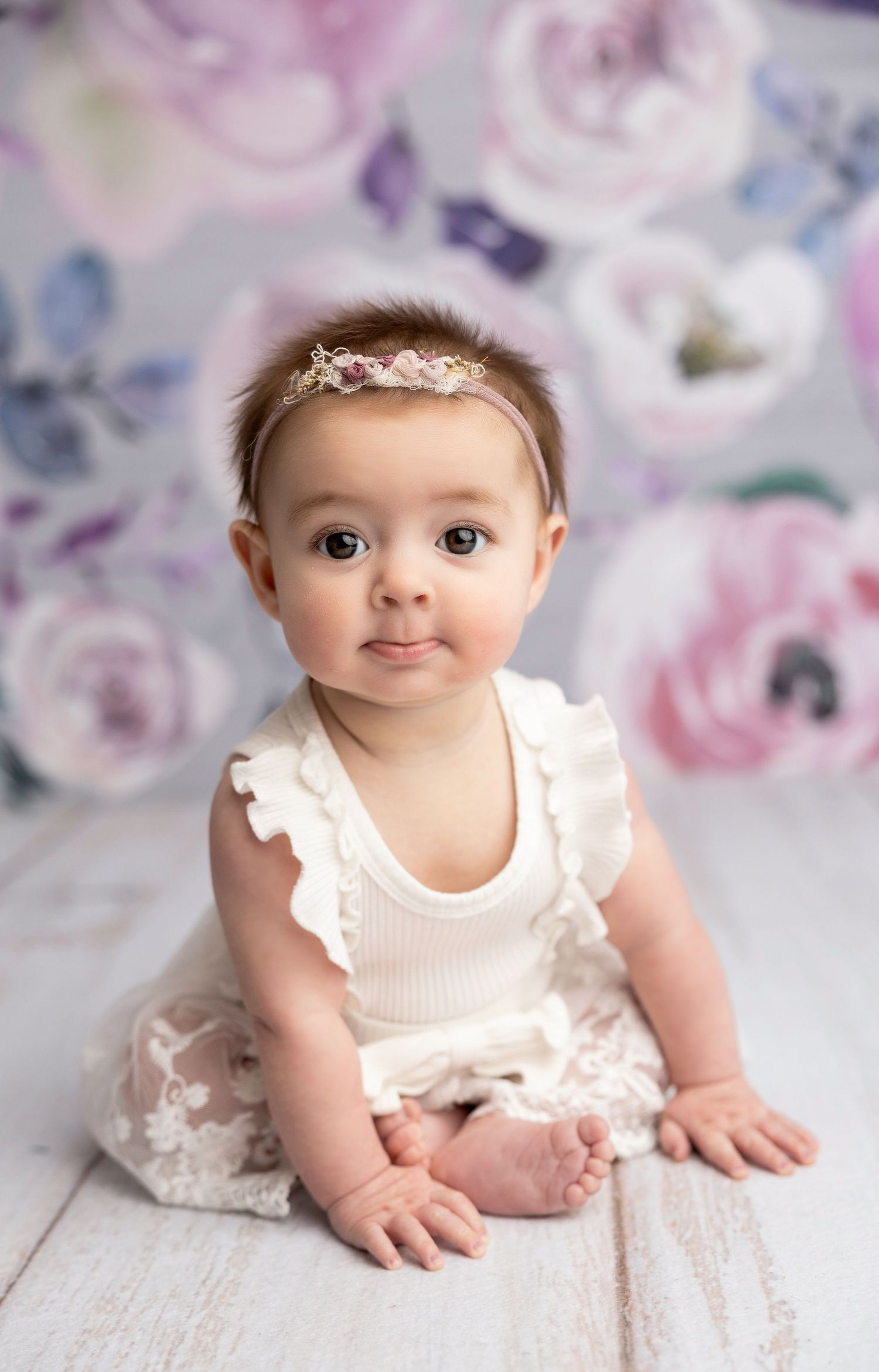 Baby in white ruffled outfit crawling on white floor with floral backdrop.