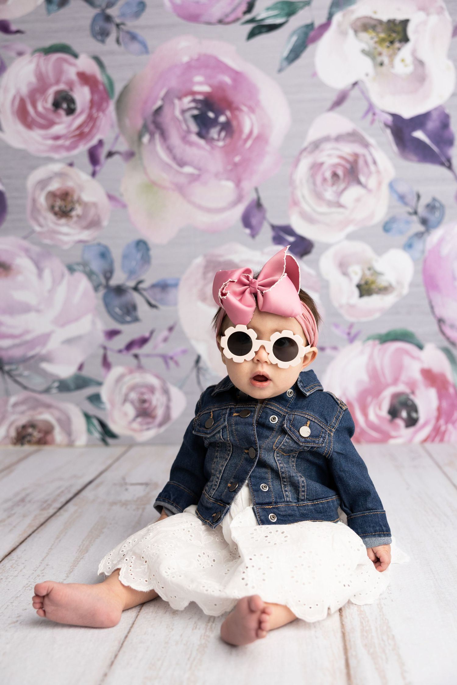 Baby in denim jacket and white dress wearing sunglasses against floral backdrop.