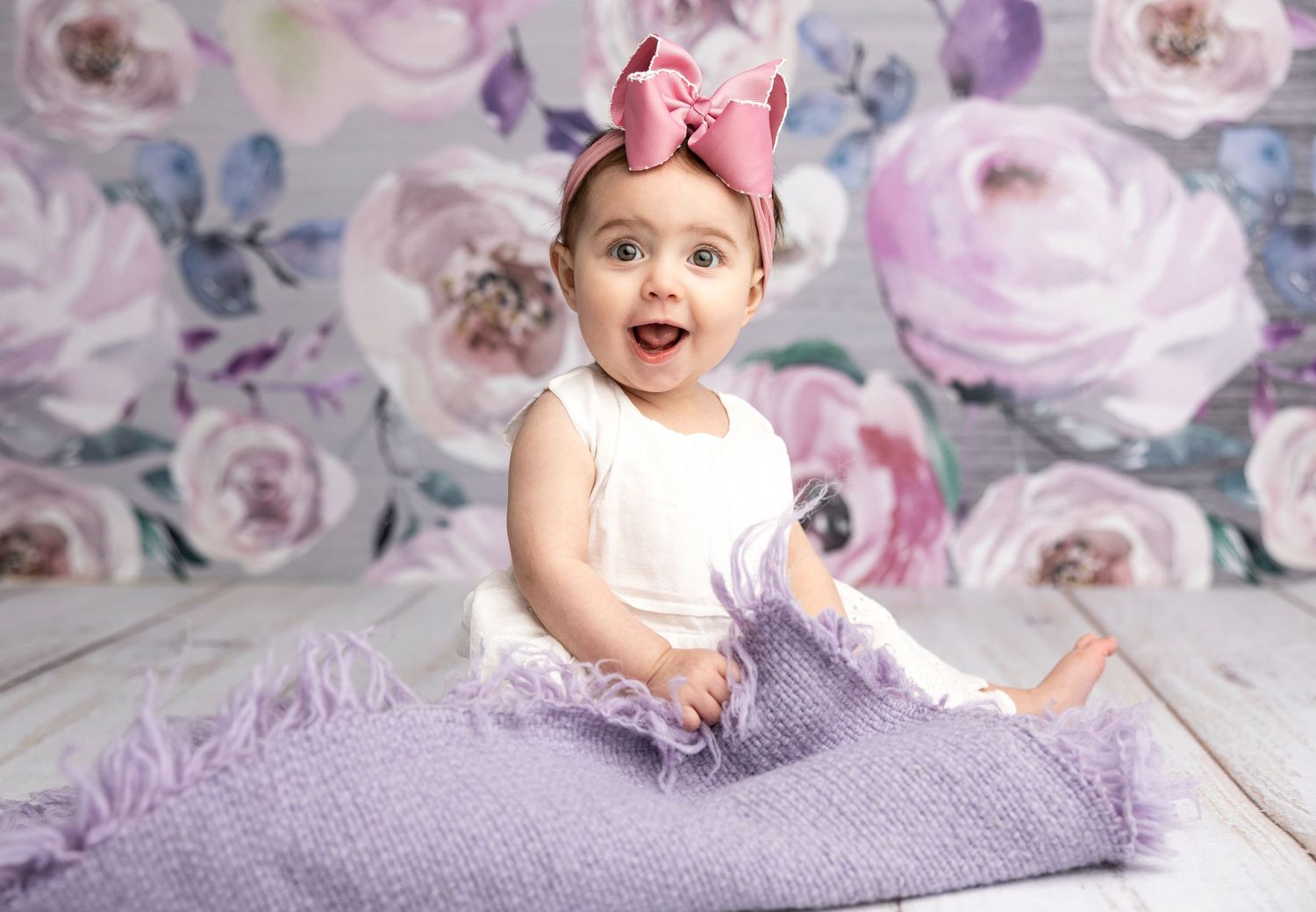 Baby wearing pink bow crawling on purple blanket with floral background.