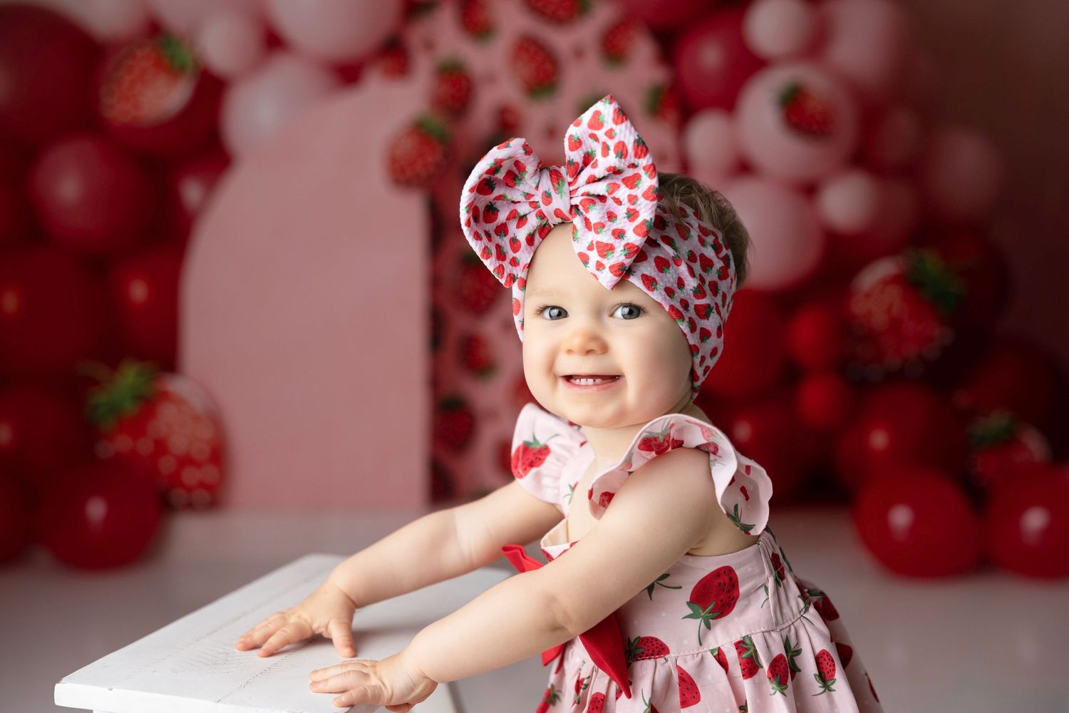 Baby in strawberry-themed outfit and headband poses against red balloon backdrop for first birthday photos.