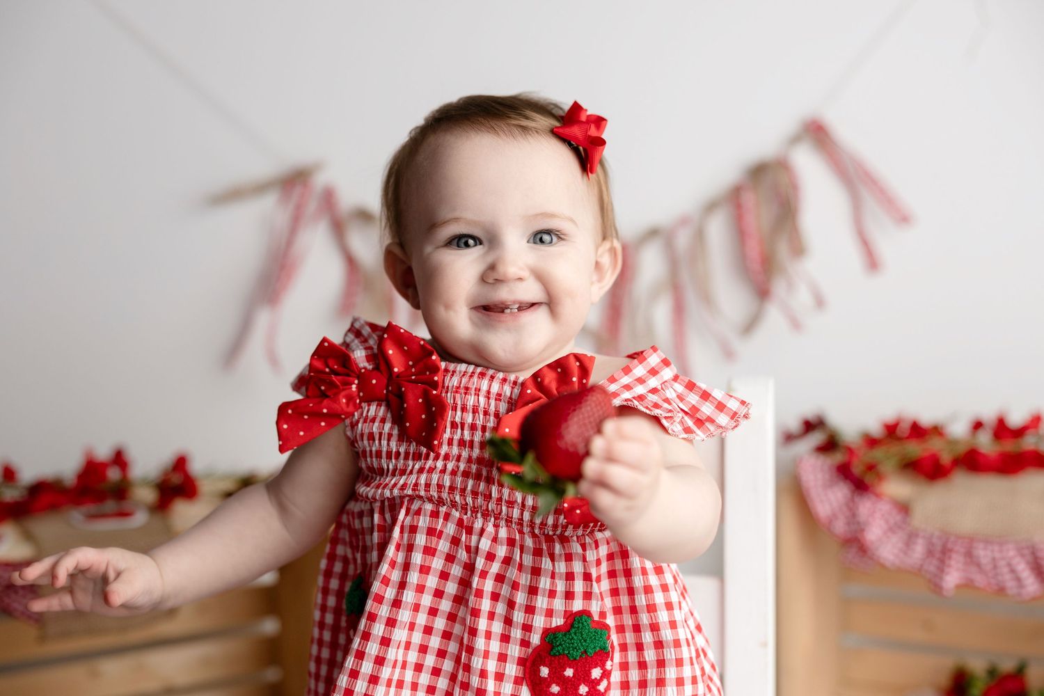 Baby girl in red gingham dress holding strawberry, smiling at camera with festive strawberry banner backdrop.