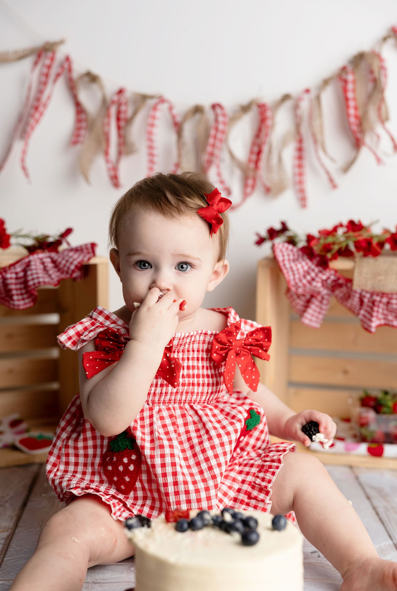Baby girl in red gingham dress eating cake at a strawberry-themed first birthday cake smash photo session.