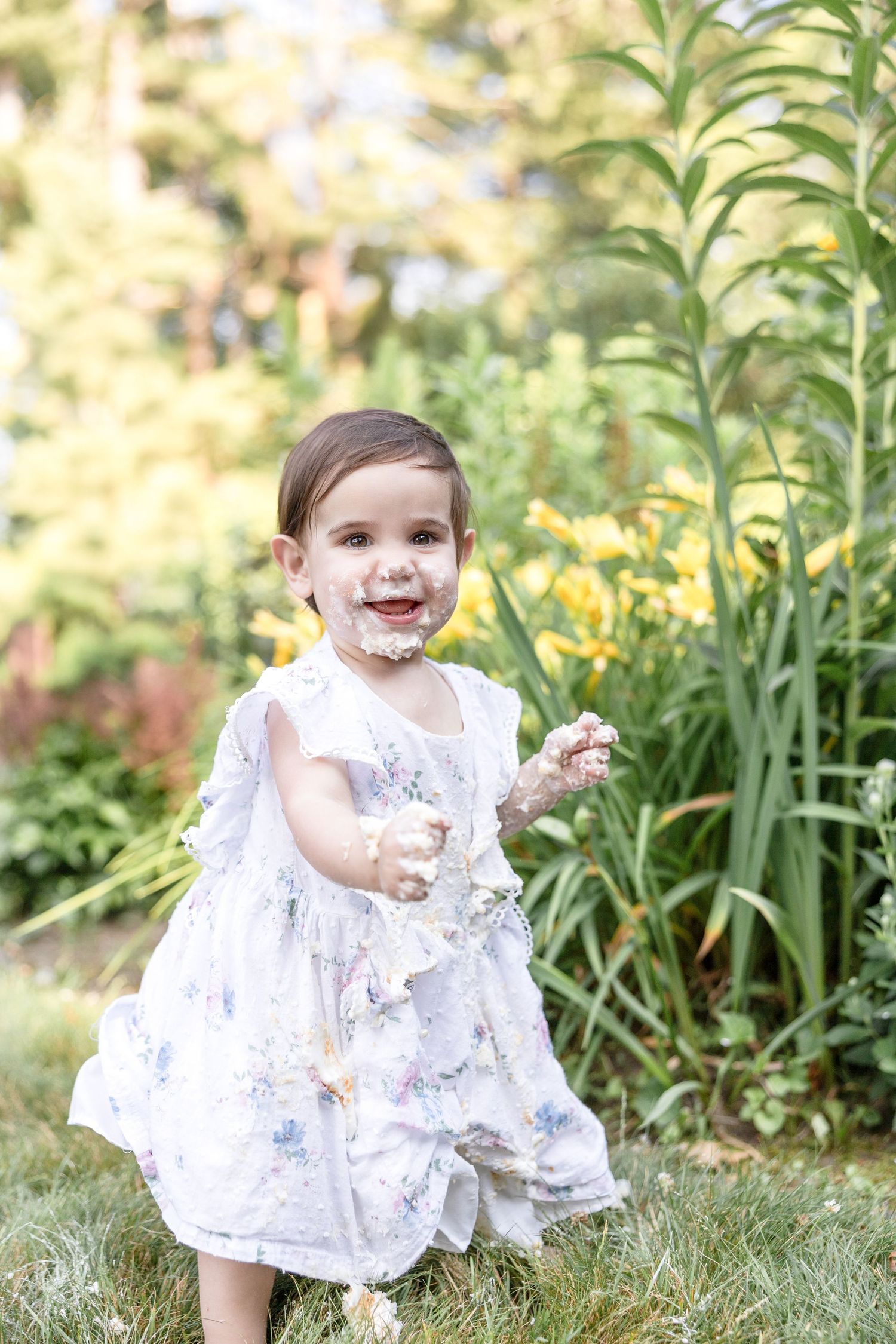 A baby in a white floral dress plays in a garden setting.