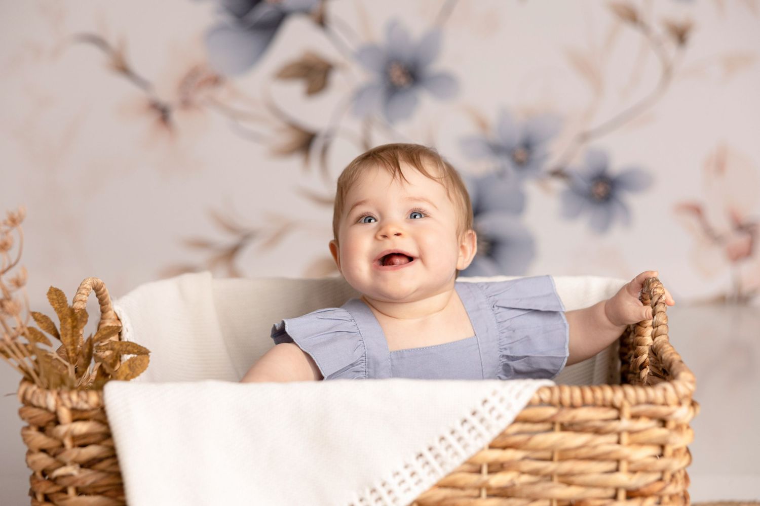 Happy baby sitting in wicker basket against floral backdrop.